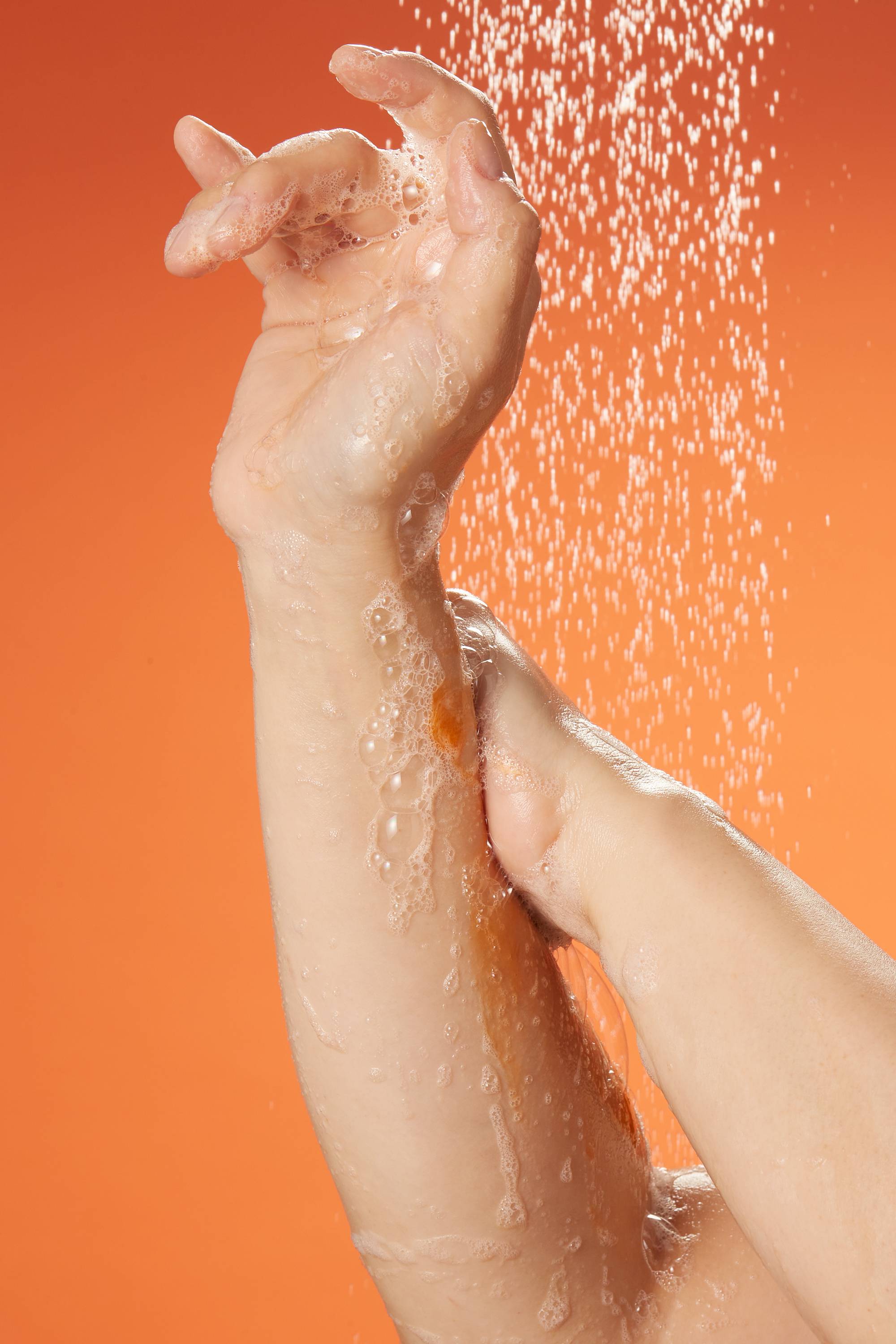 A close-up of the model's hands and forearms as they get soapy, shower gel suds over their skin under the shower water.
