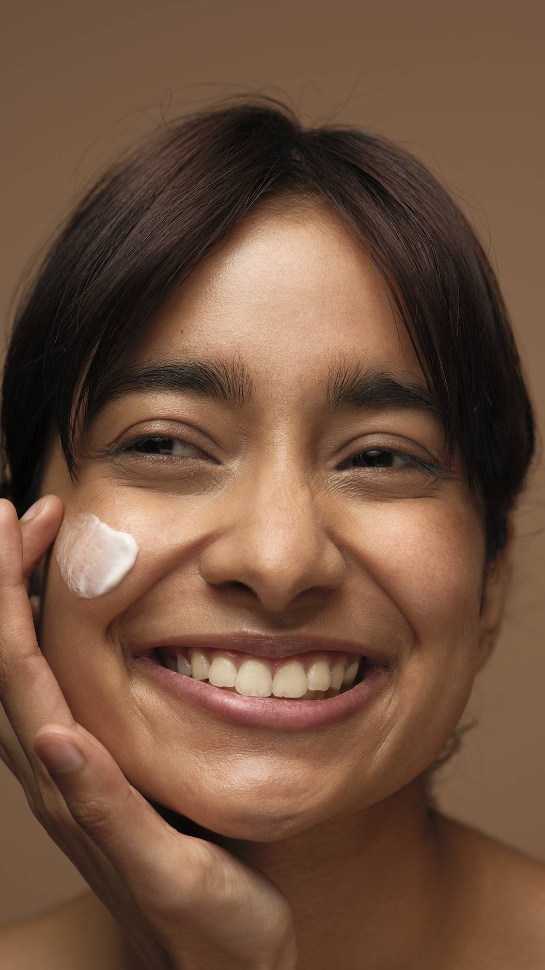 A close-up image of the model's smiling face as they start to apply the Celestial self-preserving moisturiser to one cheek. They are on a warm, earthy-brown background. 