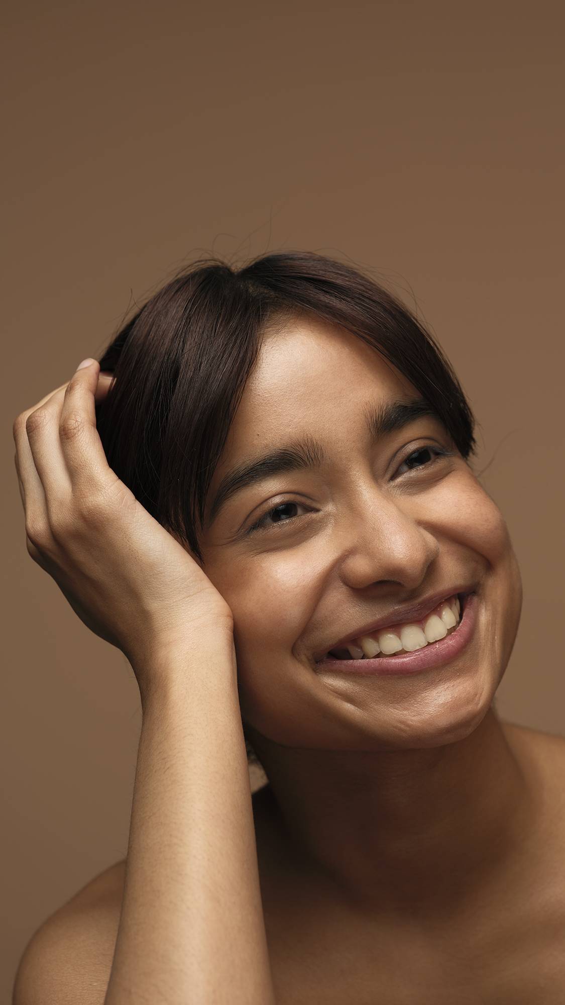 The model is on a warm, earthy-brown background with their head rested upon their hand as they are smiling with glowing, freshly moisturised skin. 