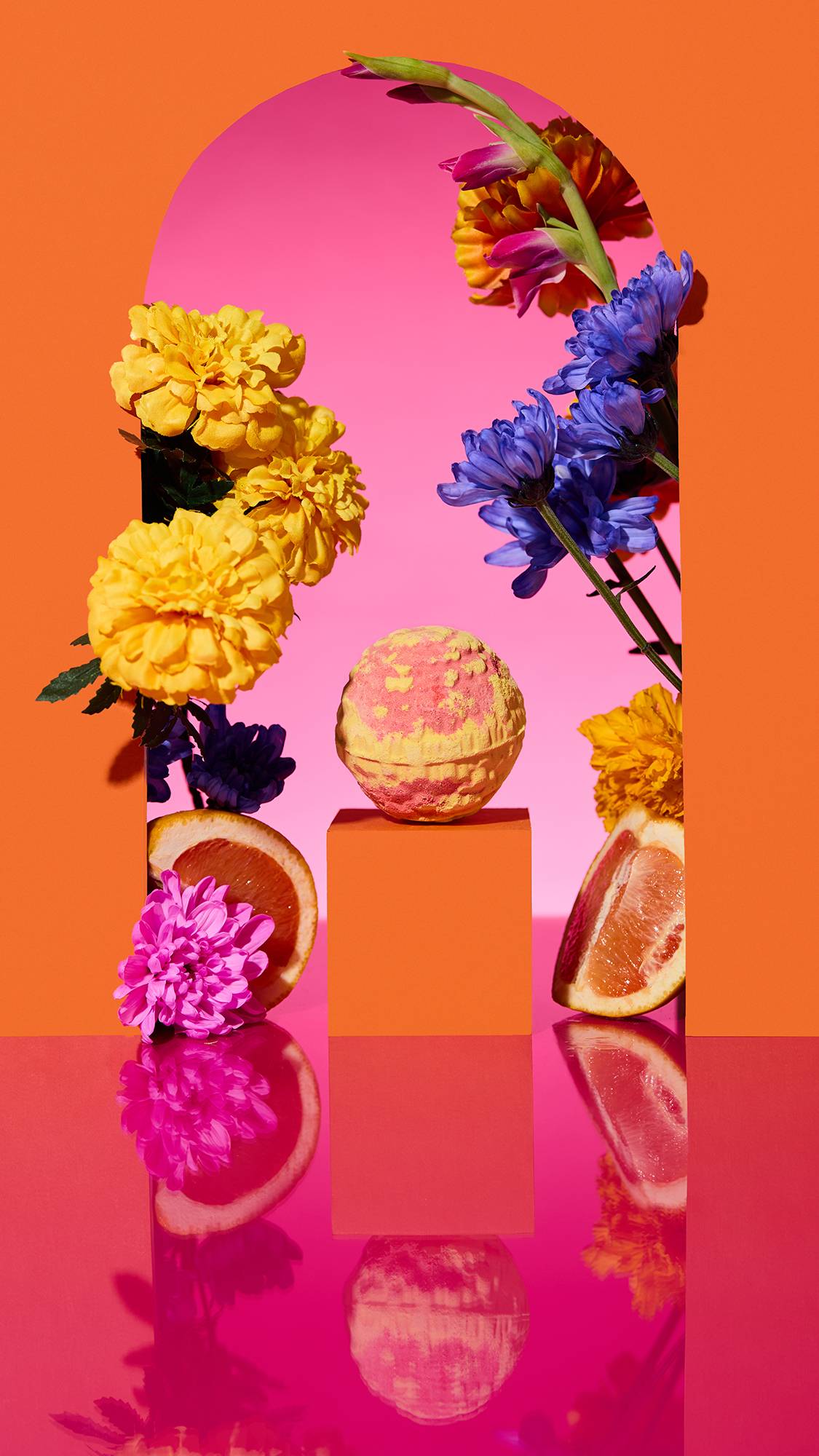 The image shows a marbled red and yellow, Dìa De Muertos-themed Cempasúchil bath bomb on a plinth, framed by orange arches on a pink background. Sliced citrus, colourful flowers, and a pink reflective surface surround the product in a vibrant scene.