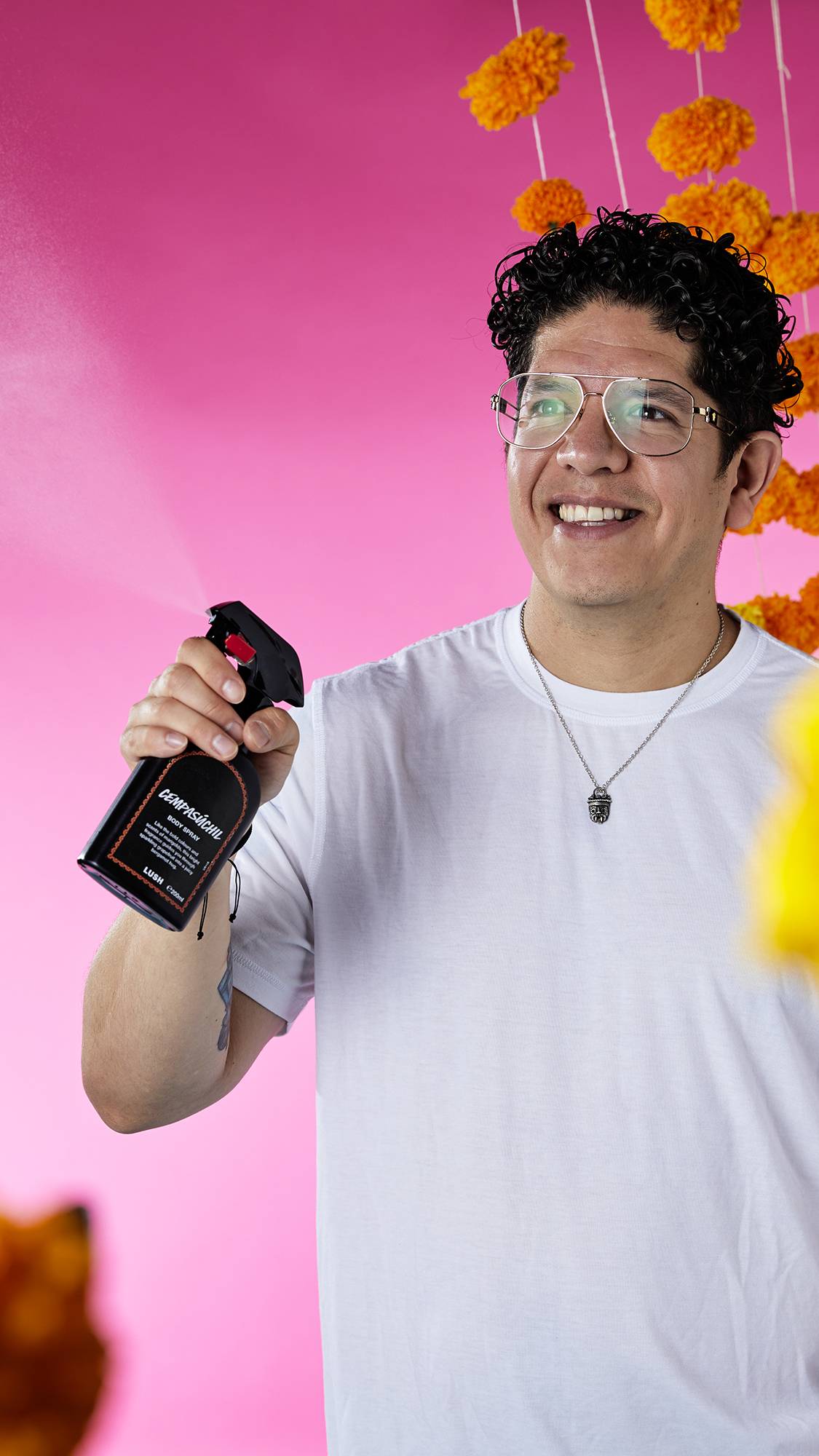 The image shows a person with dark hair, smiling as they spray a bottle of Cempasúchil Body Spray. A fine mist of product is visible against a vibrant pink background with hanging marigold garlands.