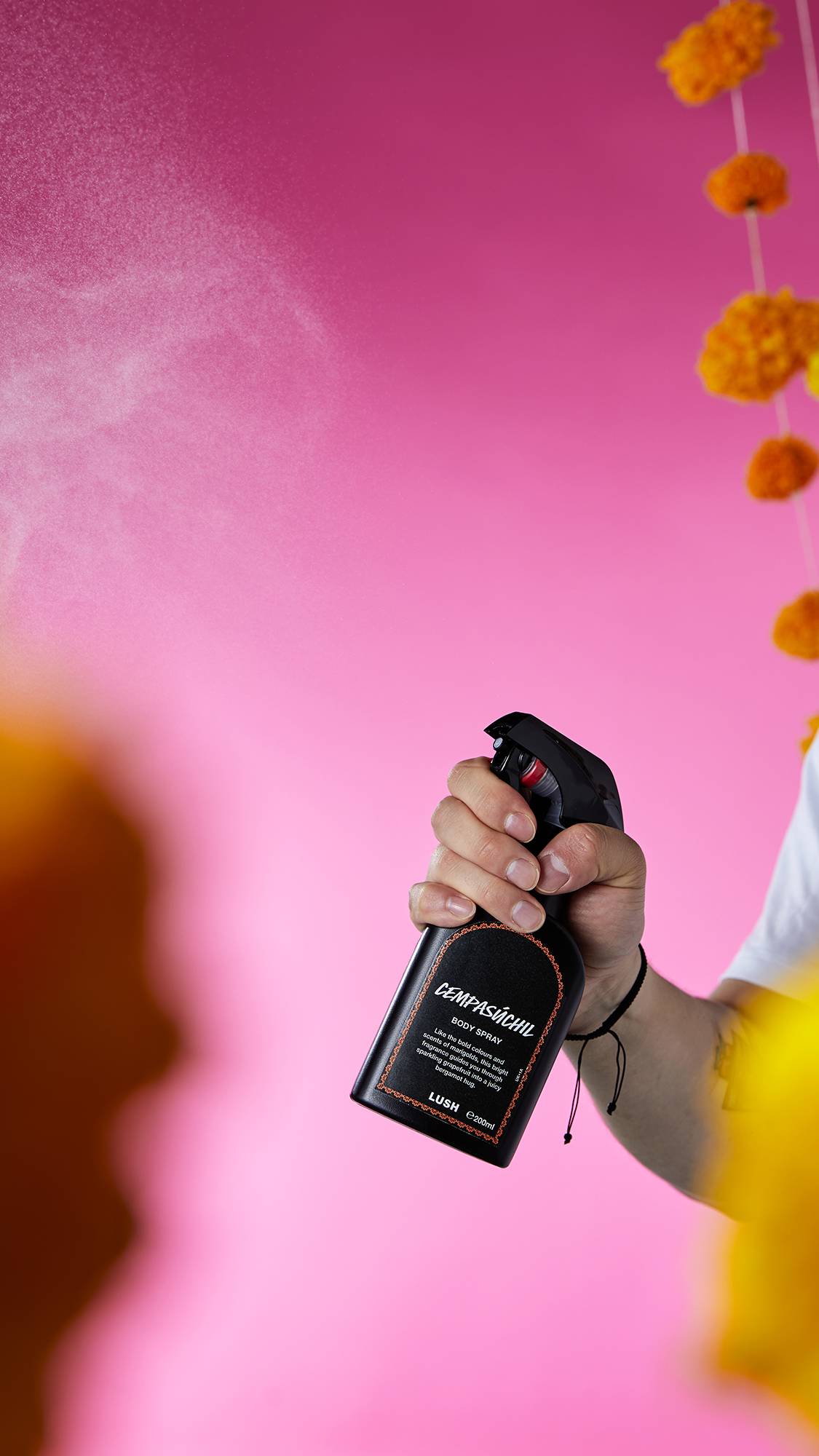 The image shows a hand spraying a bottle of Cempasúchil Body Spray. A fine mist of product is visible against a vibrant pink background with hanging marigold garlands.