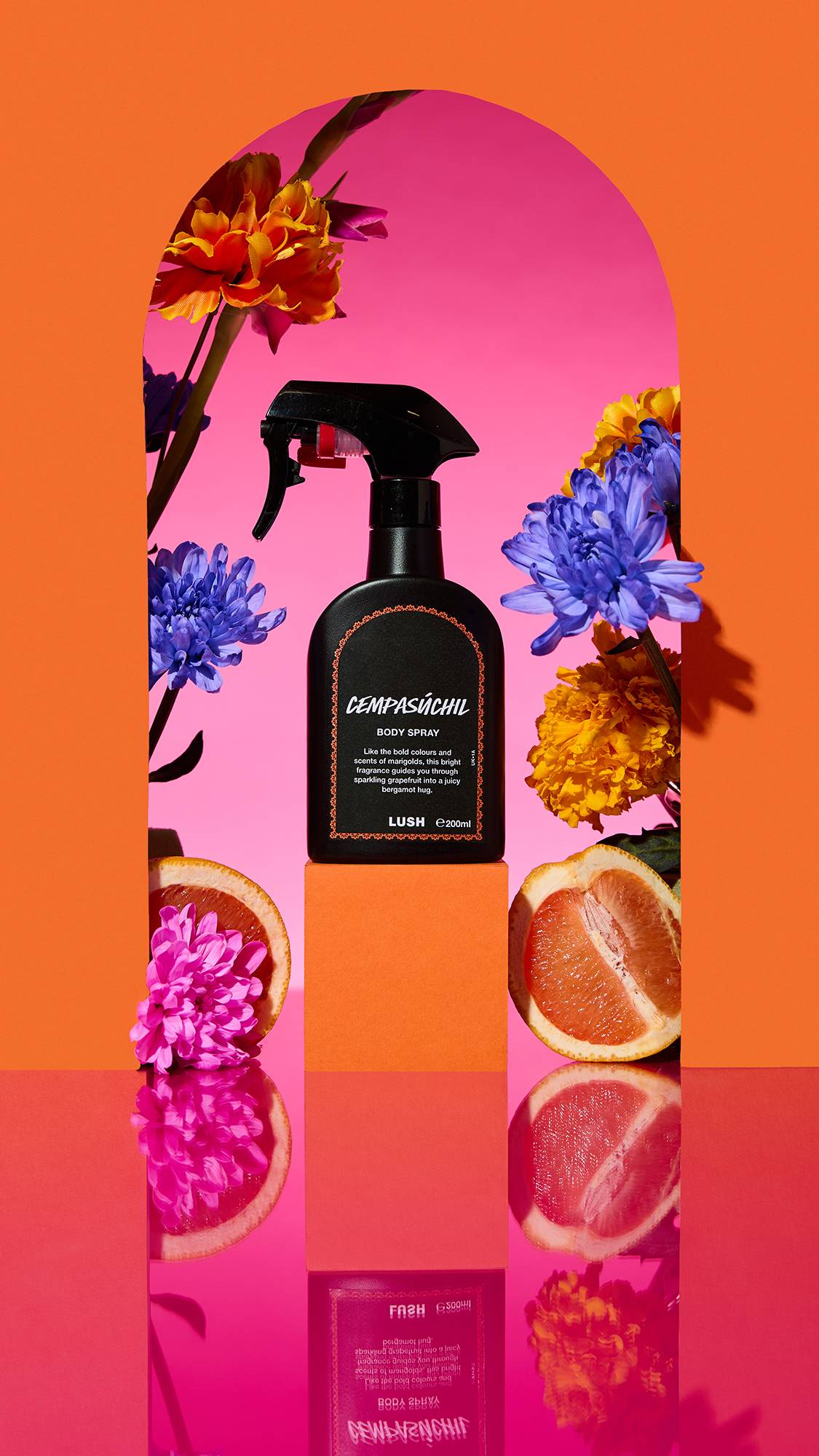 The image shows a black spray bottle with an orange-bordered label of the Cempasúchil body spray on a plinth. A colourful arch frames the scene, with sliced citrus, flowers, and a cinnamon stick surrounding the product on a reflective pink surface.