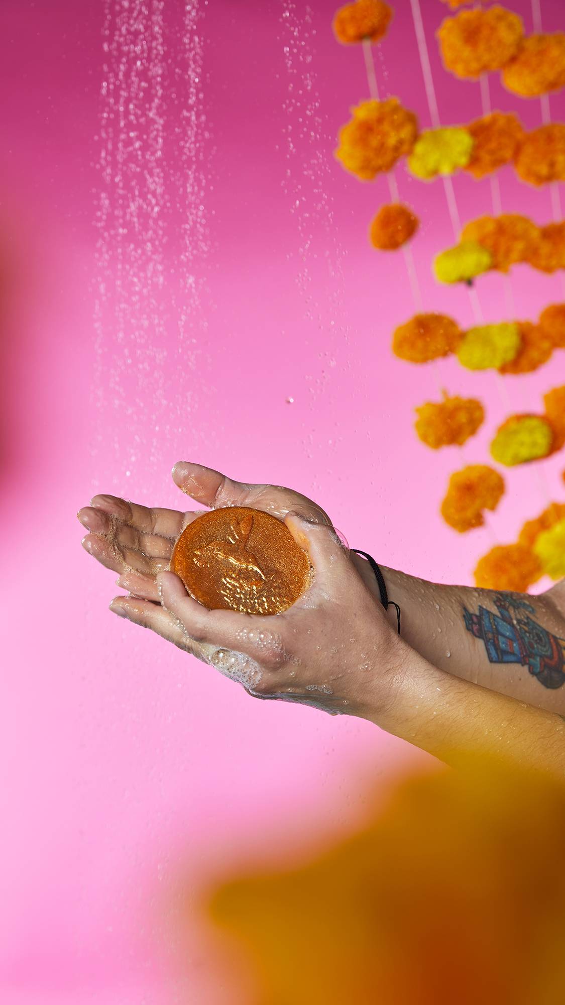 Cempasúchil Soap. The image shows a pair of hands holding a round, golden soap with a shimmering hummingbird embossed on top. Bubbly lather and water droplets gather in the cupped palms, against a vibrant pink background.
