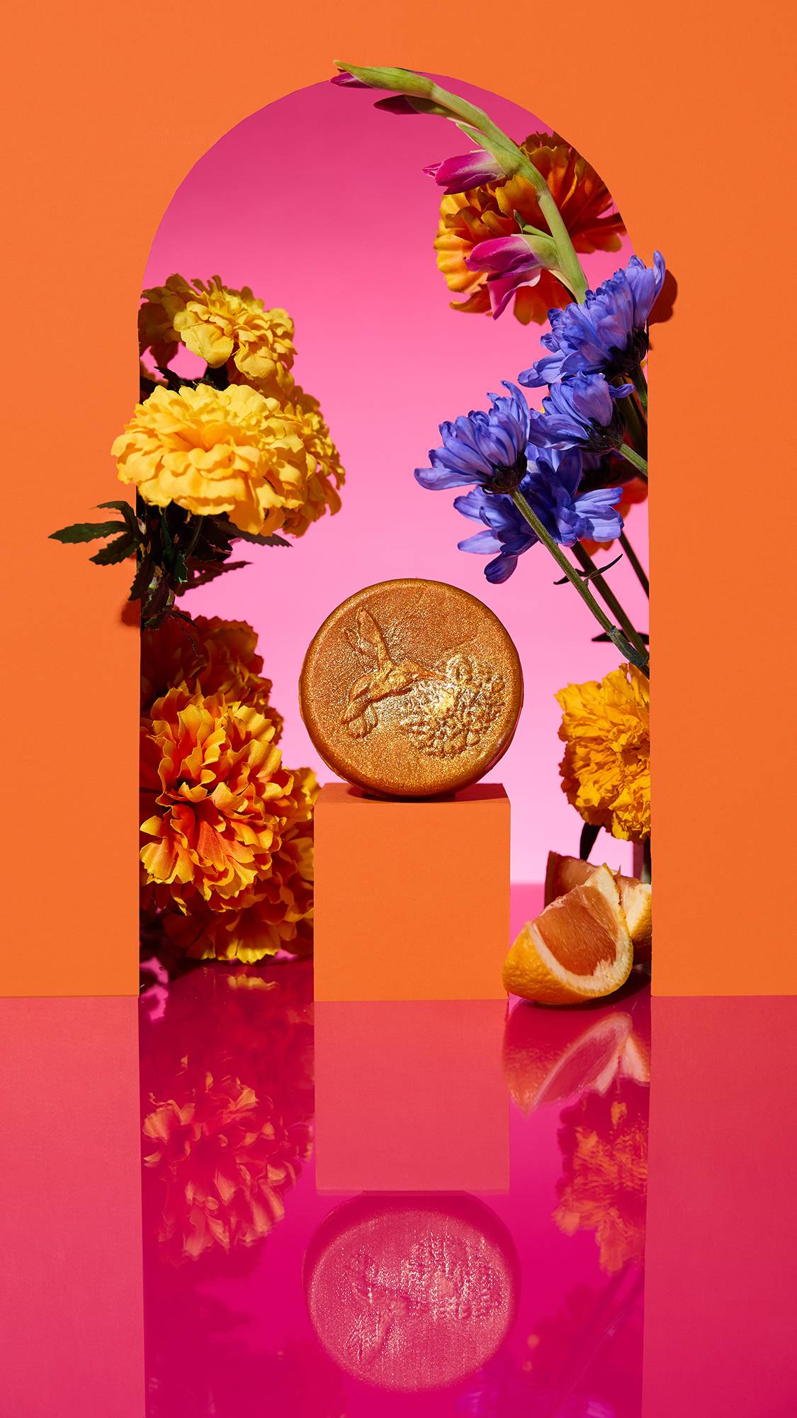 The image shows the circular, golden, Cempasúchil Soap on a plinth. A colourful arch frames the scene, with sliced citrus, flowers, and a cinnamon stick surrounding the product on a reflective pink surface.