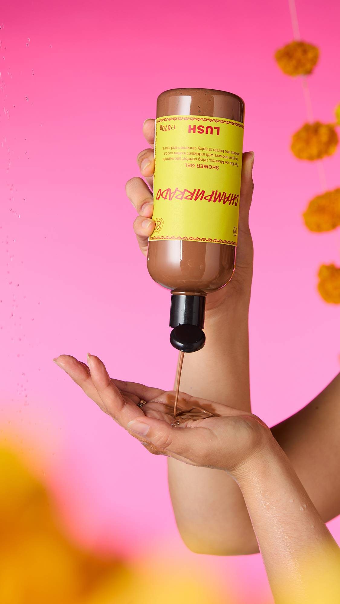 The image shows hands holding an upside-down bottle of caramel-coloured Champurrado Shower Gel. A thick stream pours into a cupped palm, with water droplets and marigold garlands against a vibrant pink background.