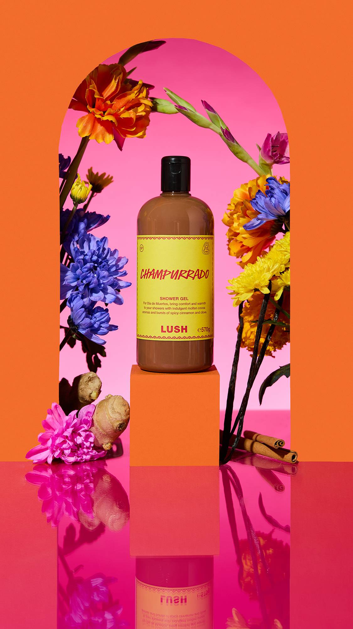 The image shows a clear bottle with a yellow label of the caramel-coloured Champurrado shower gel on a plinth. A colourful arch frames the scene, with sliced citrus, flowers, and a cinnamon stick surrounding the product on a reflective pink surface.