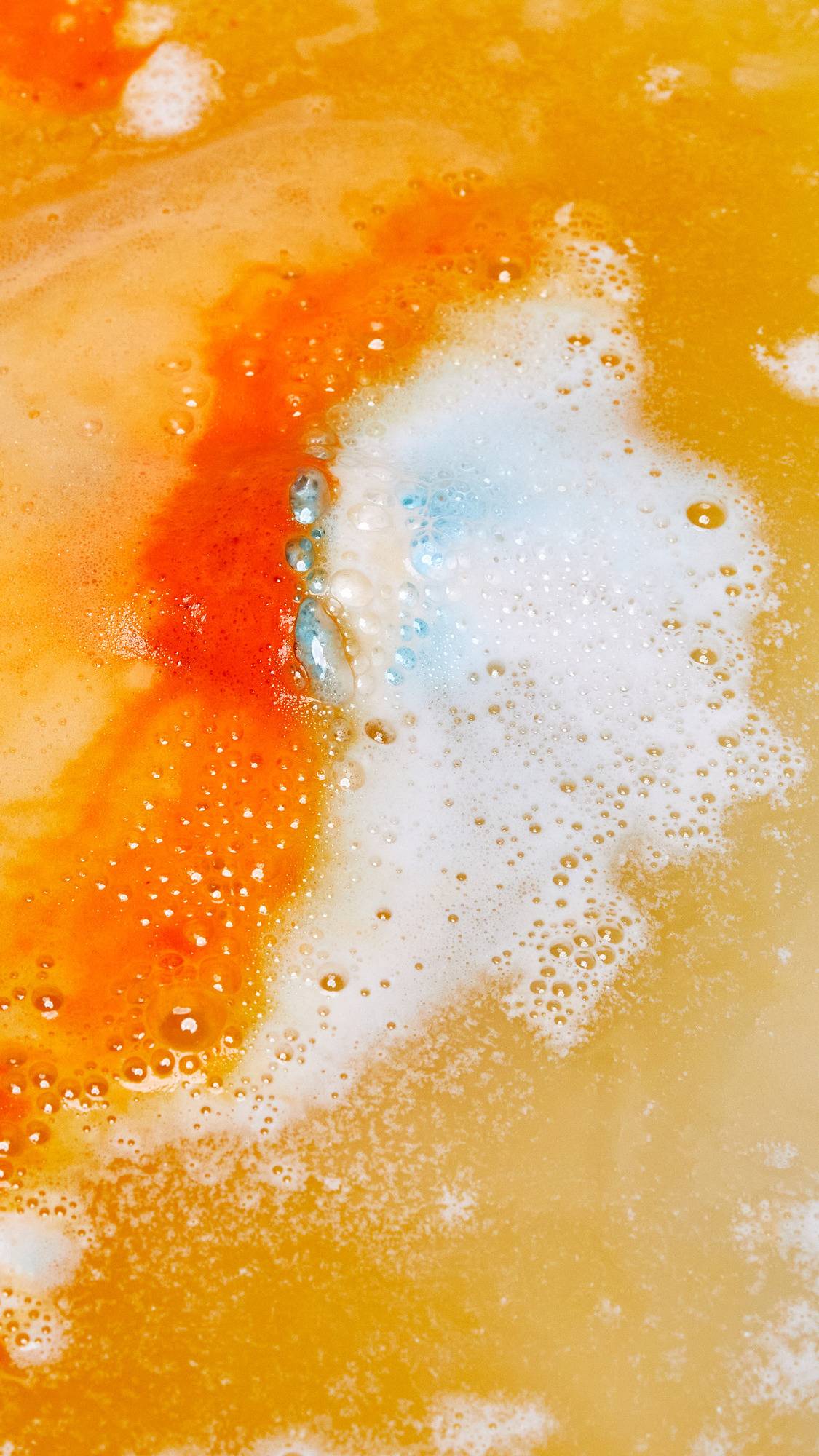 The Chelsea Morning bath bomb has just been placed into the bath water. The top half gives off vibrant, orange, foamy ripples while the bottom of the bath bomb releases delicate blue and white foamy swirls.