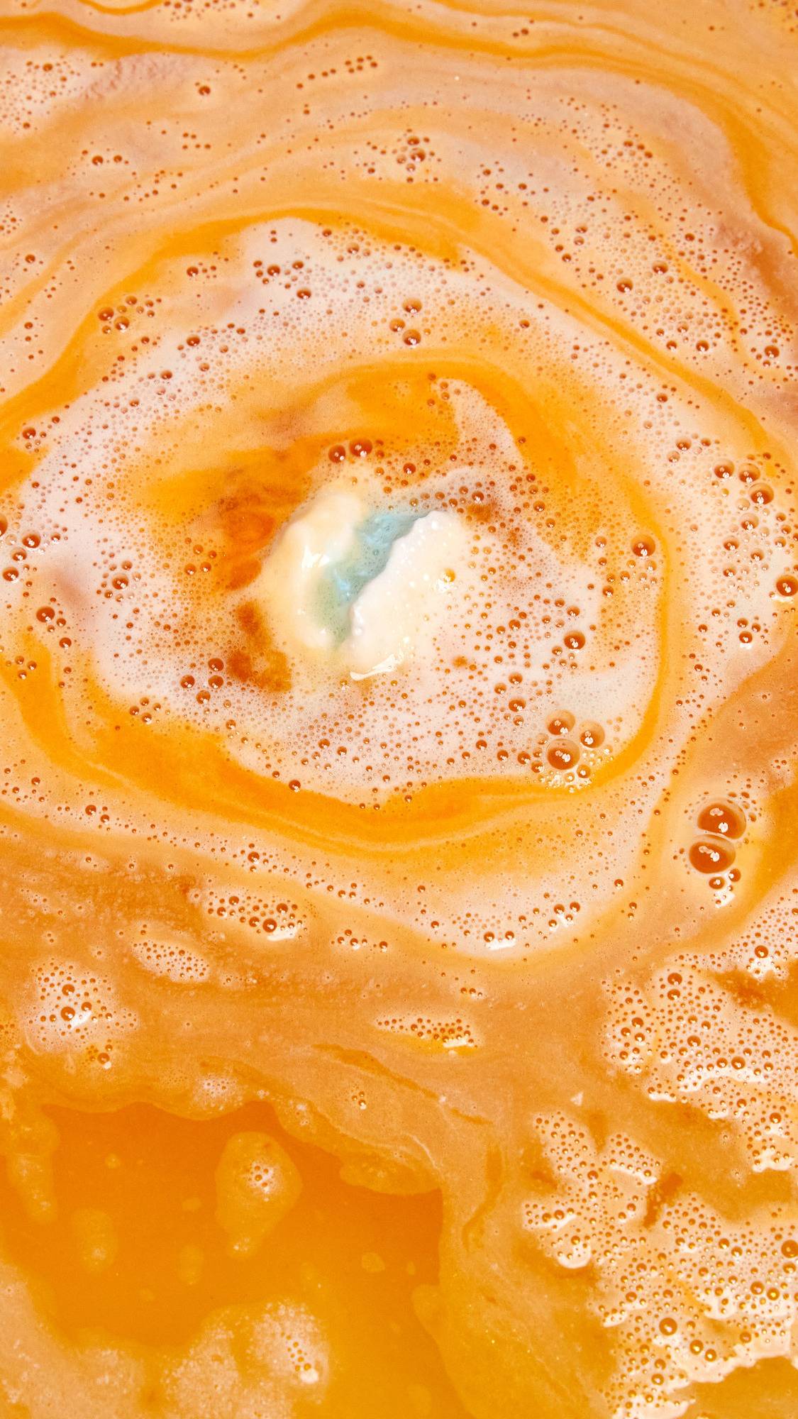 The Chelsea Morning bath bomb is dissolving in the bath water creating alternating layers of foamy, white and bright orange ripples.
