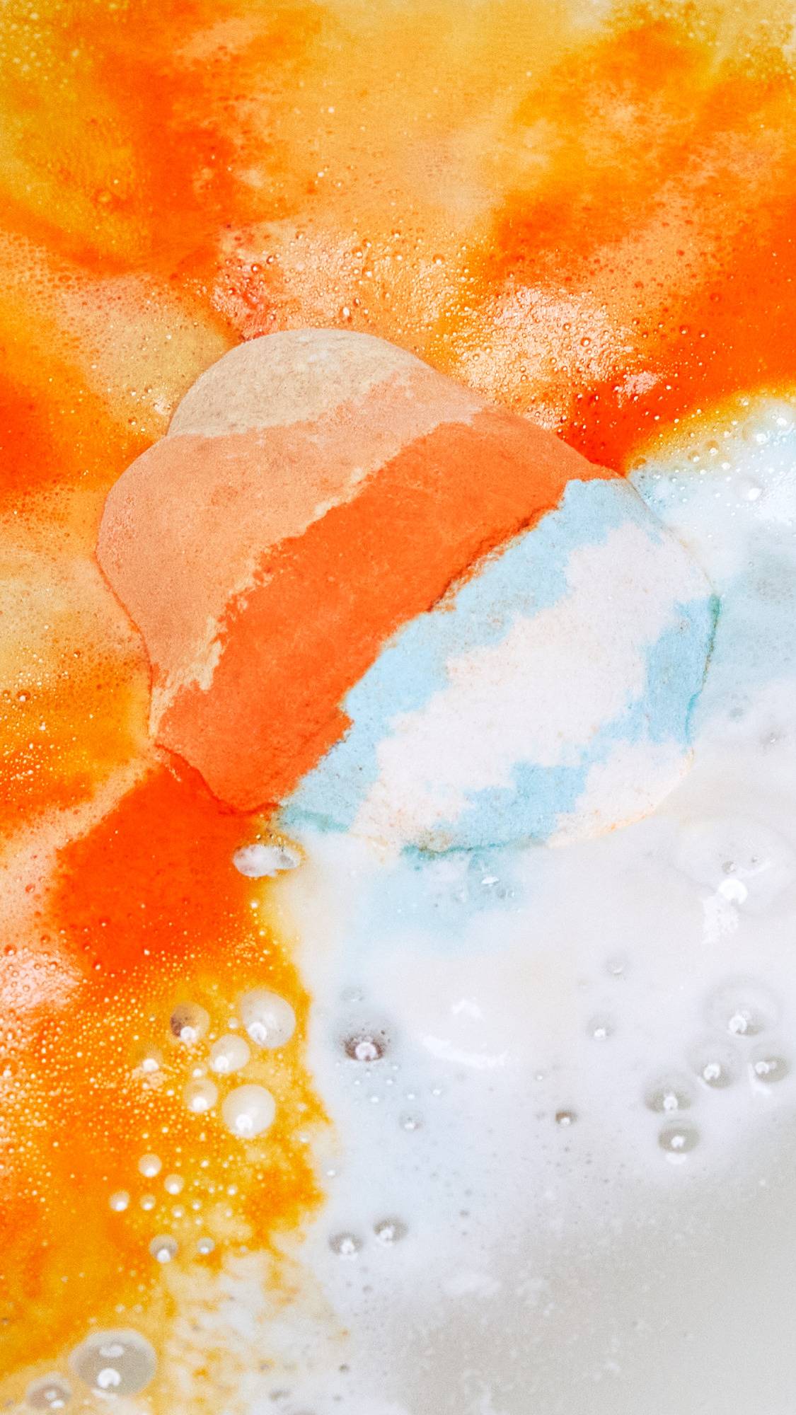 The Chelsea Morning bath bomb has just been placed into the bath water. The top half gives off vibrant, orange, foamy ripples while the bottom of the bath bomb releases delicate blue and white foamy swirls.
