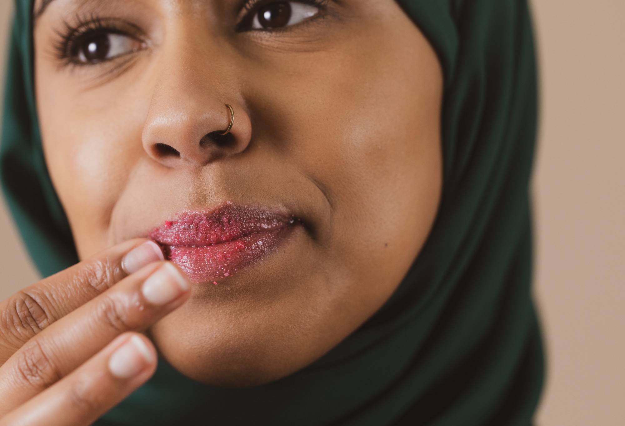 A person with a nose piercing, wearing a Hijab, scrubs their pouted lips with  fuchsia pink Cherry sugar lip scrub.