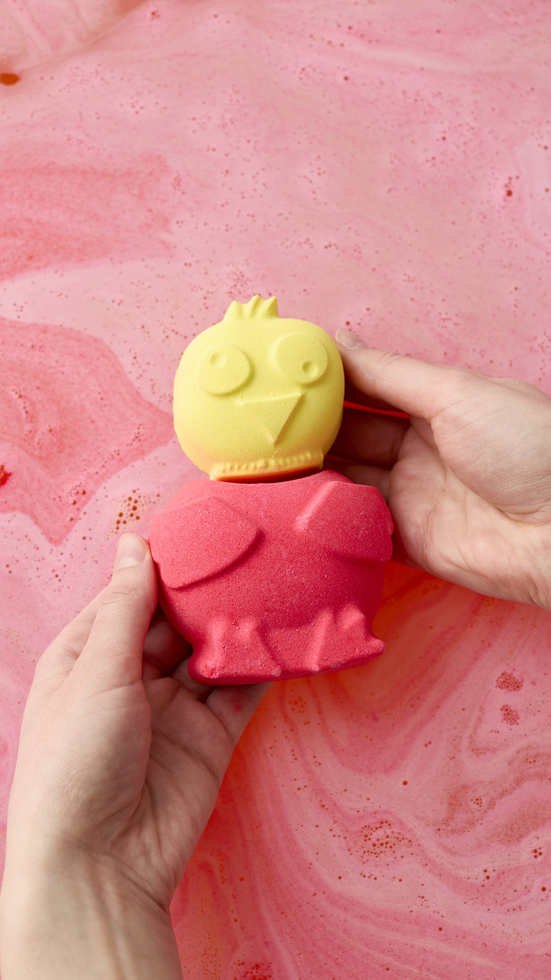 A yellow and pink Chirpy Chick bath bomb is held by two hands over vibrant pink bathwater with frothy bubbles. The bath bomb has a yellow chick head with wide eyes and a pink bird-shaped body.