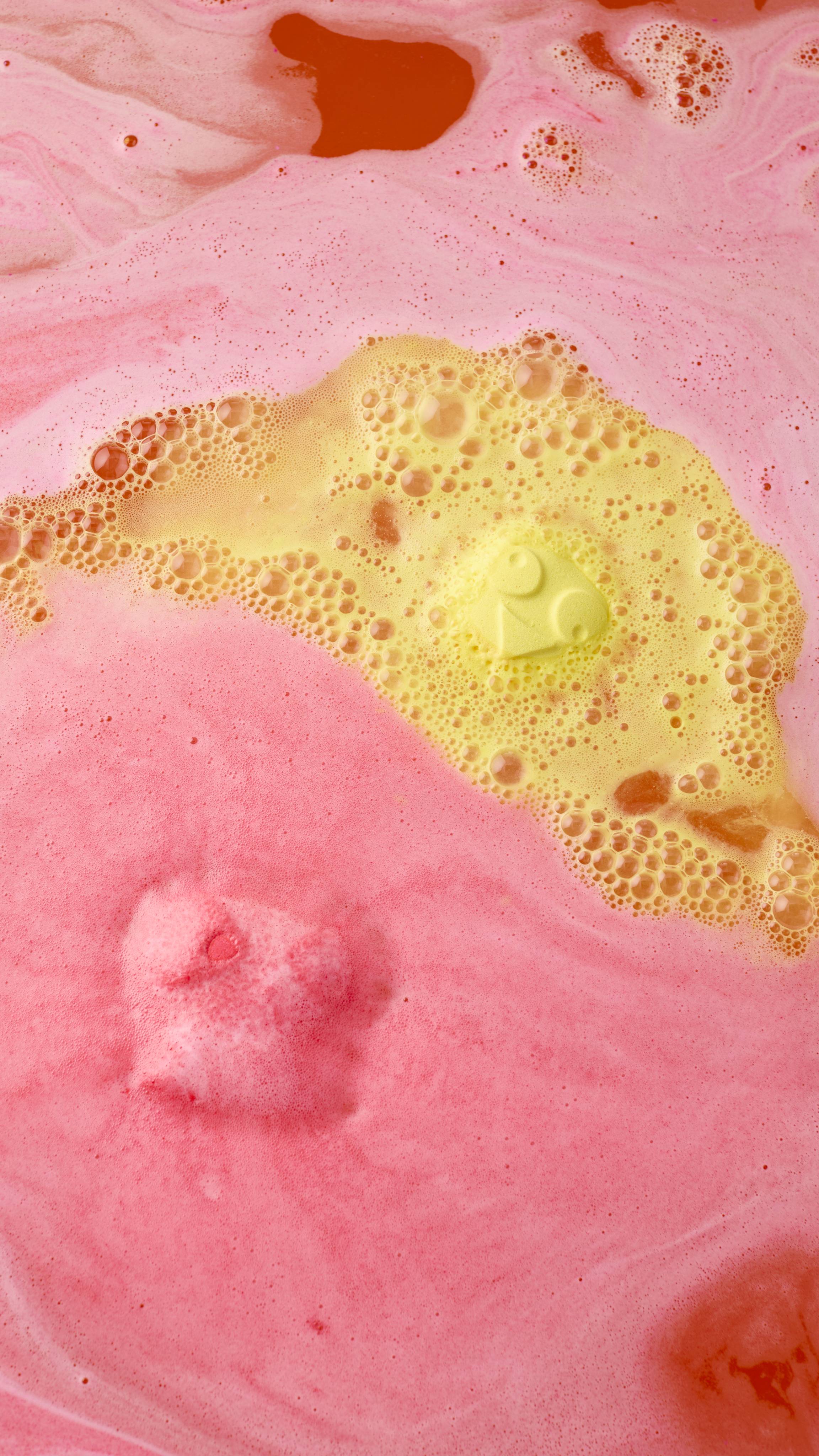 The Chirpy Chick bath bomb dissolves in bathwater, creating a vibrant landscape of thick pink and yellow foam. The yellow chick face is still visible amidst the bubbling, colorful swirls. All glitter used is plastic-free.