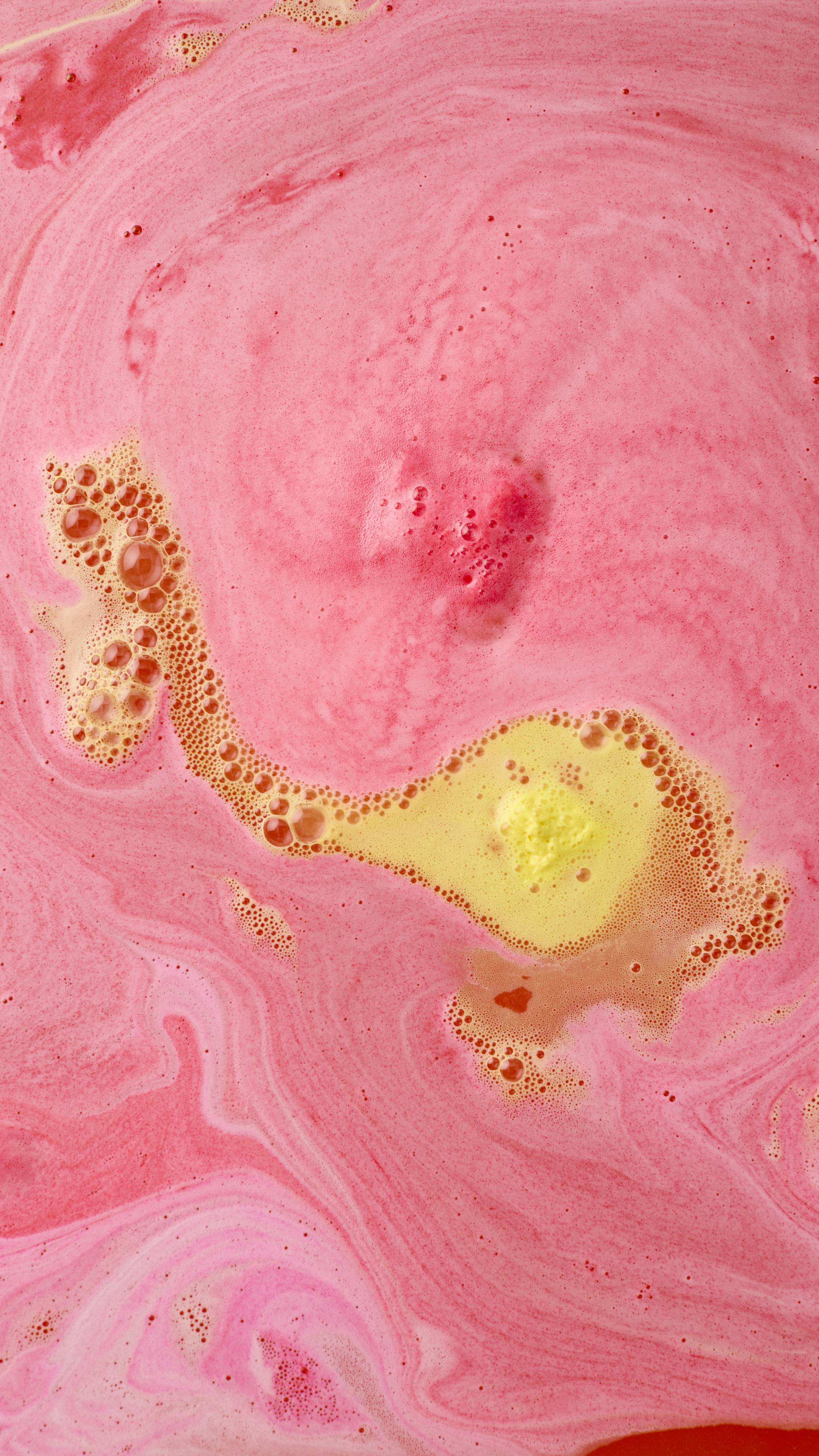 The Chirpy Chick bath bomb dissolves into a sea of vibrant pink water, releasing a trail of thick yellow foam across the surface. The colors swirl together to create a bright, marbled pattern of pink, orange, and yellow.