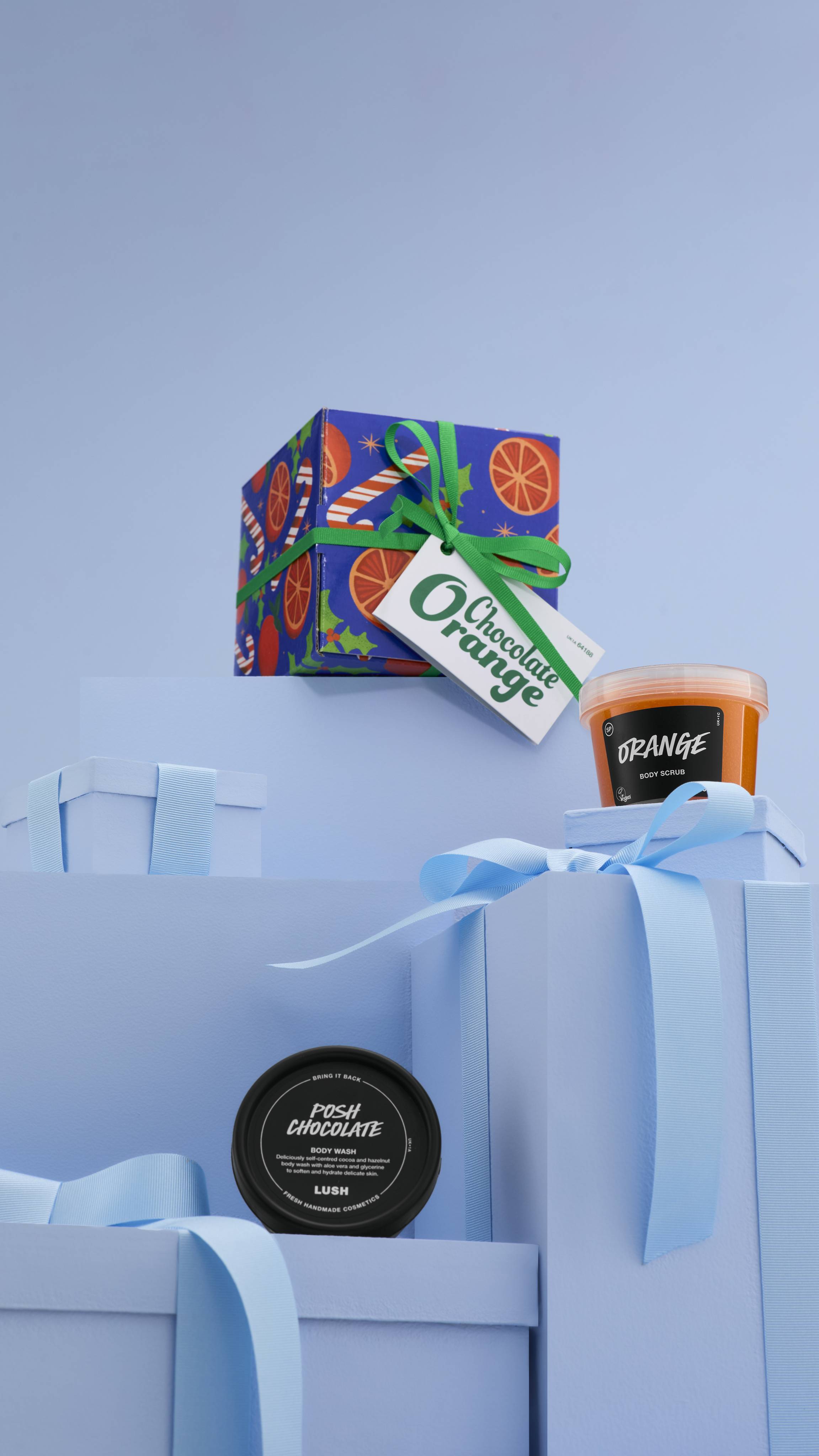 The image shows a large pile of differently-sized wrapped pale blue gifts with ribbons, on a matching blue background. The colourful Chocolate Orange gift box sits on top of the pile, and the two included potted products sit on lower blue gifts. 