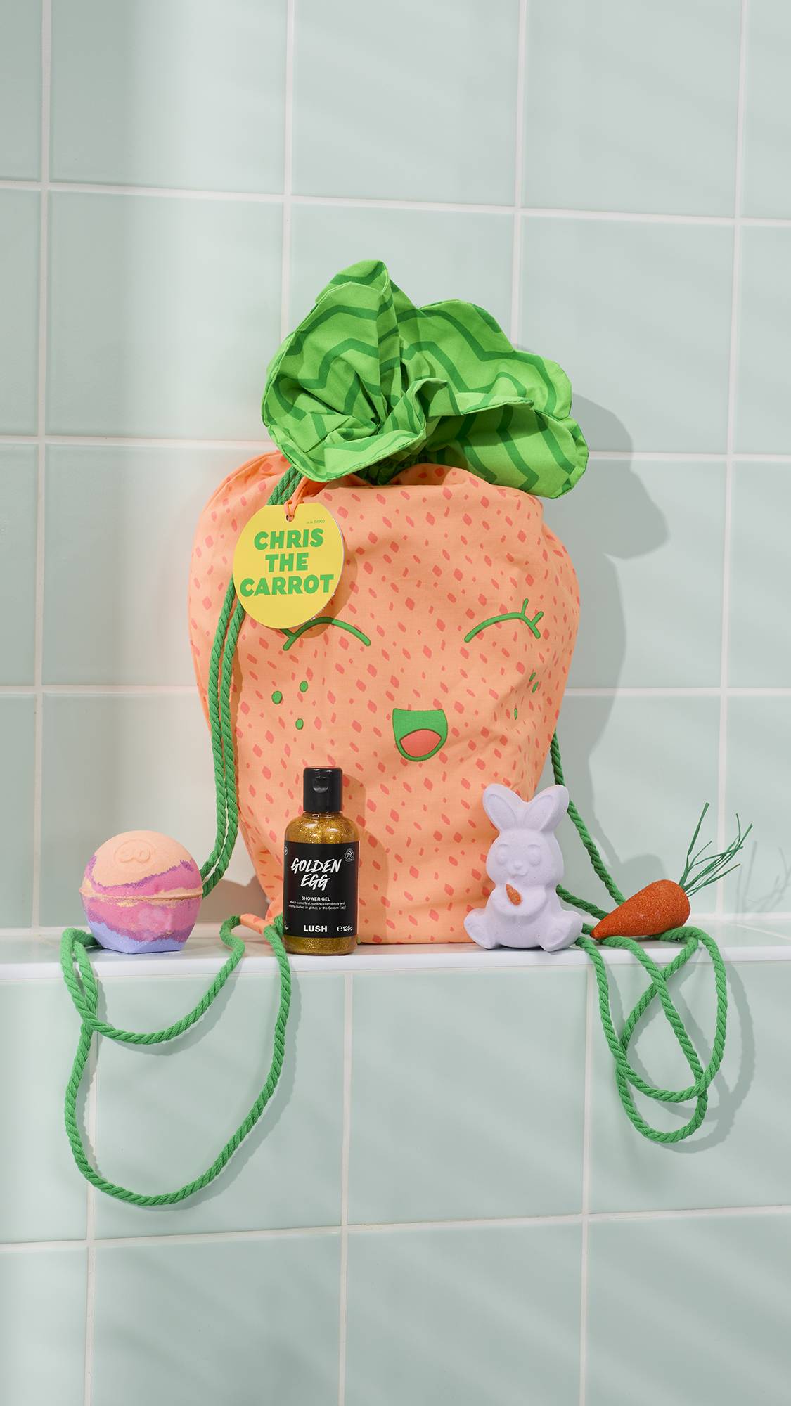 The image shows the orange "Chris The Carrot" gift drawstring bag with a green leafy top sitting on a tiled bathroom ledge alongside the included products of two bath bombs, a bubble bar and a bottle of shower gel.