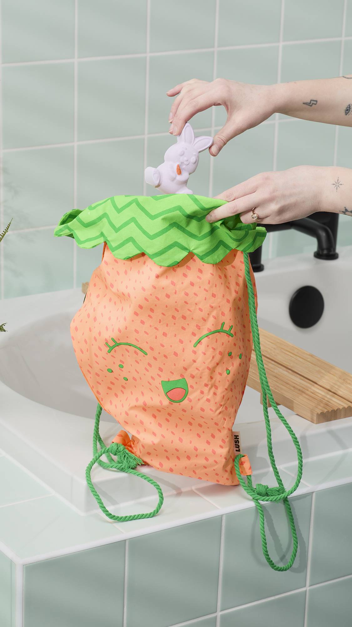 A model's tattooed hand retrieves a small, bunny-shaped bath bomb from the top of a large "Chris The Carrot" drawstring bag. The bag features a smiling face, and the scene is set against the edge of a white bathtub and mint-green wall tiles.