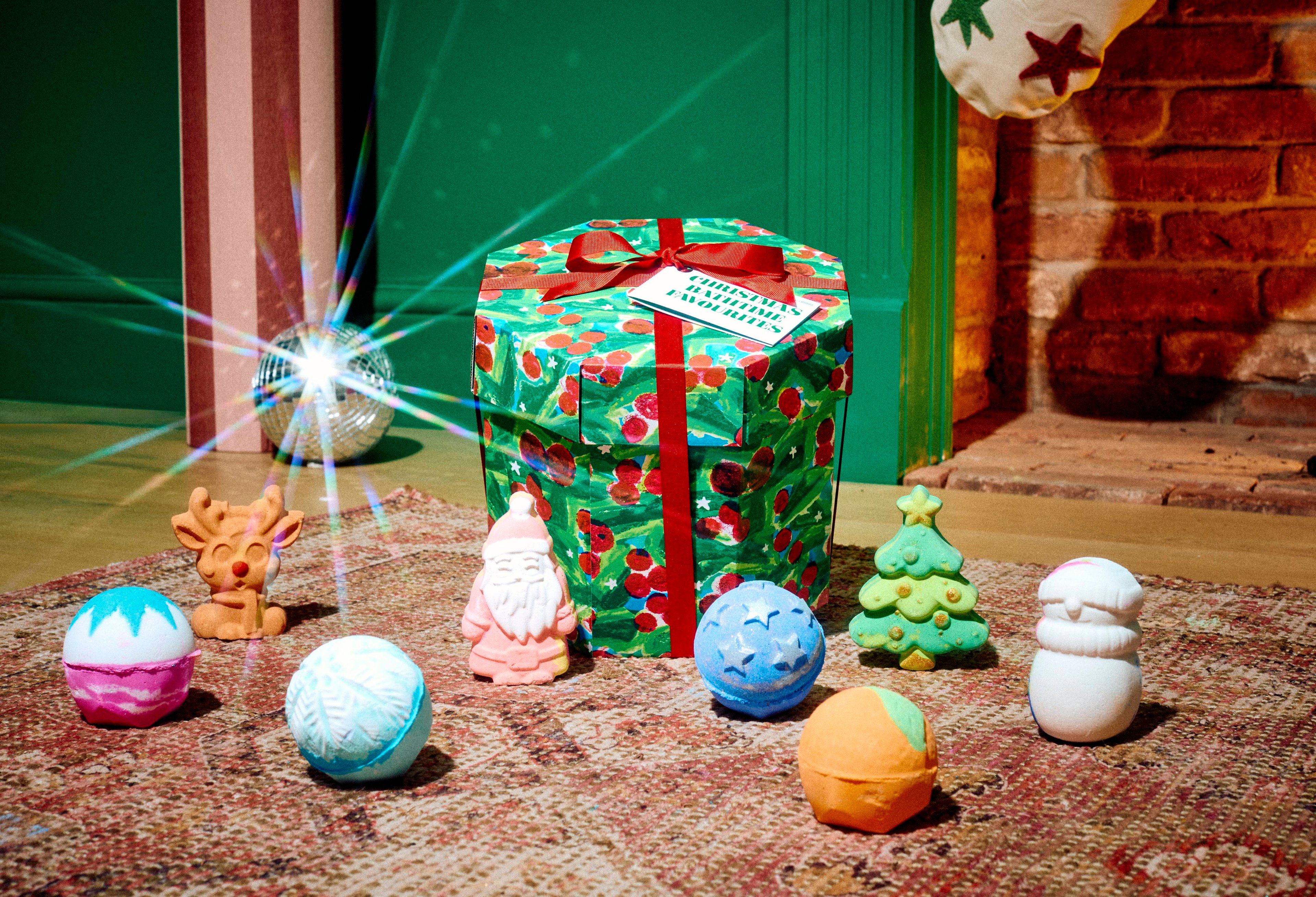 The Lush Christmas Bathtime Favourites gift set sits on a patterned rug surrounded by eight assorted festive bath products, including a snowman, reindeer, Father Christmas, Christmas tree, and spherical bath bombs, in a cosy room setting.