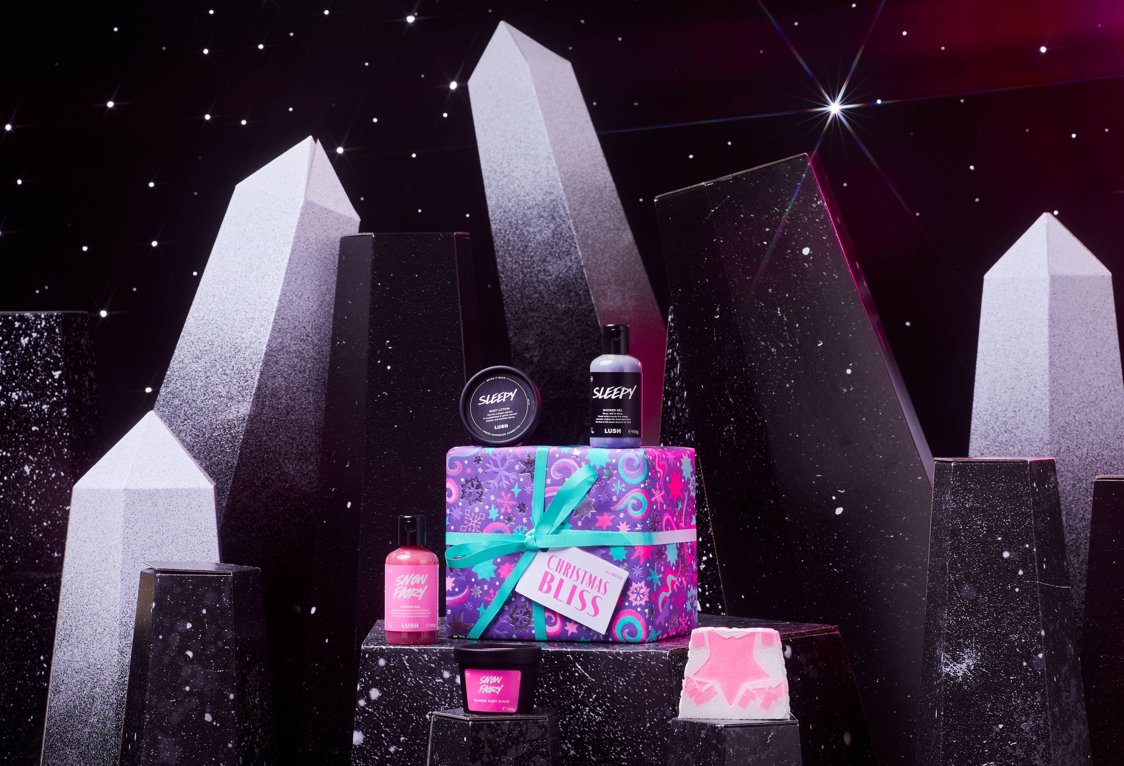 Christmas Bliss gift box and 5 products are on a dark chromatic platform among geometric shapes. 