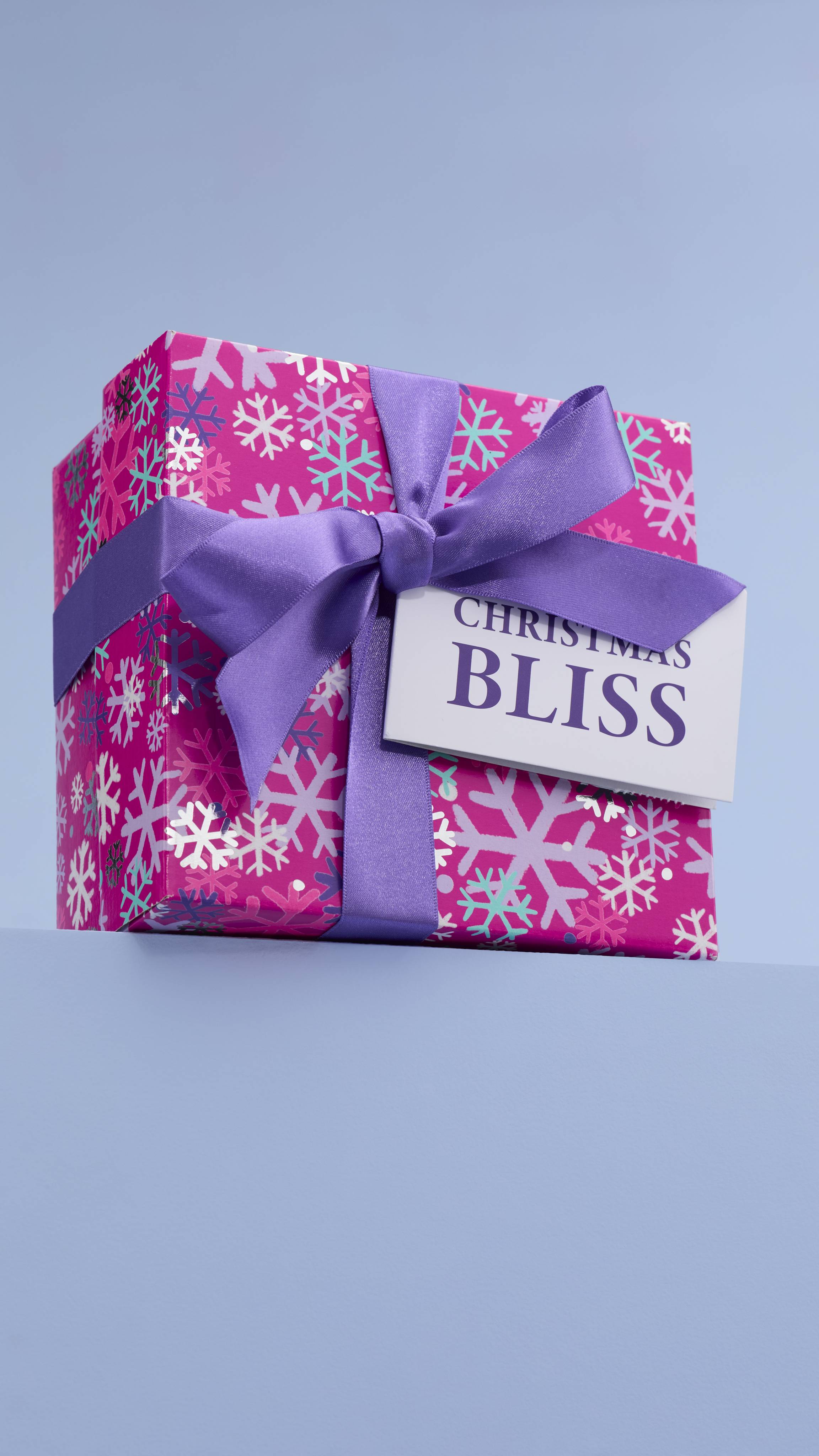  The image shows the pink, snowflake-designed "Christmas Bliss" gift box, tied with purple ribbon and a white gift tag, sitting on a pale-blue platform against a matching blue background. 