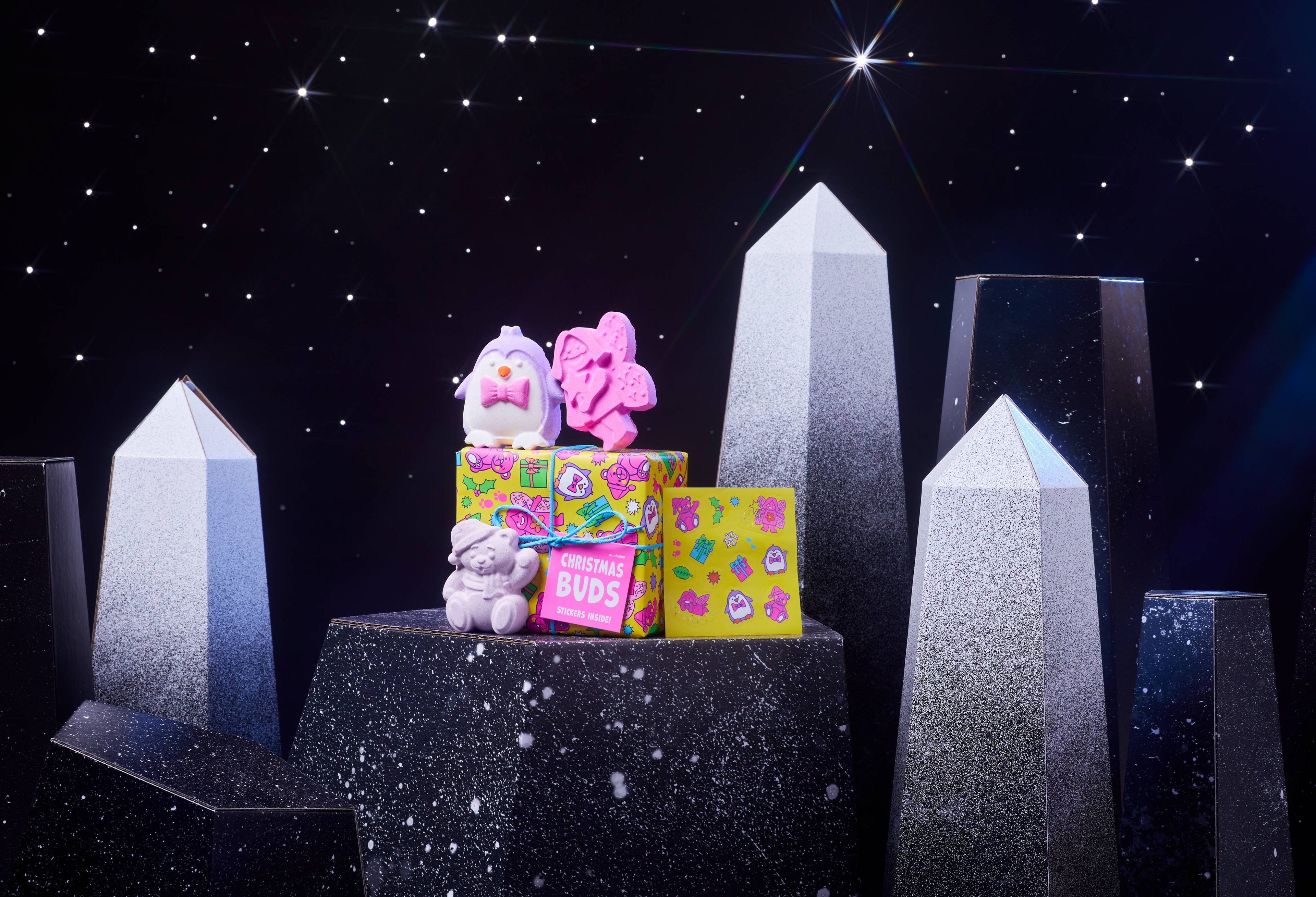 Christmas Buds gift box and three bath bombs are on a dark chromatic platform among geometric shapes. 