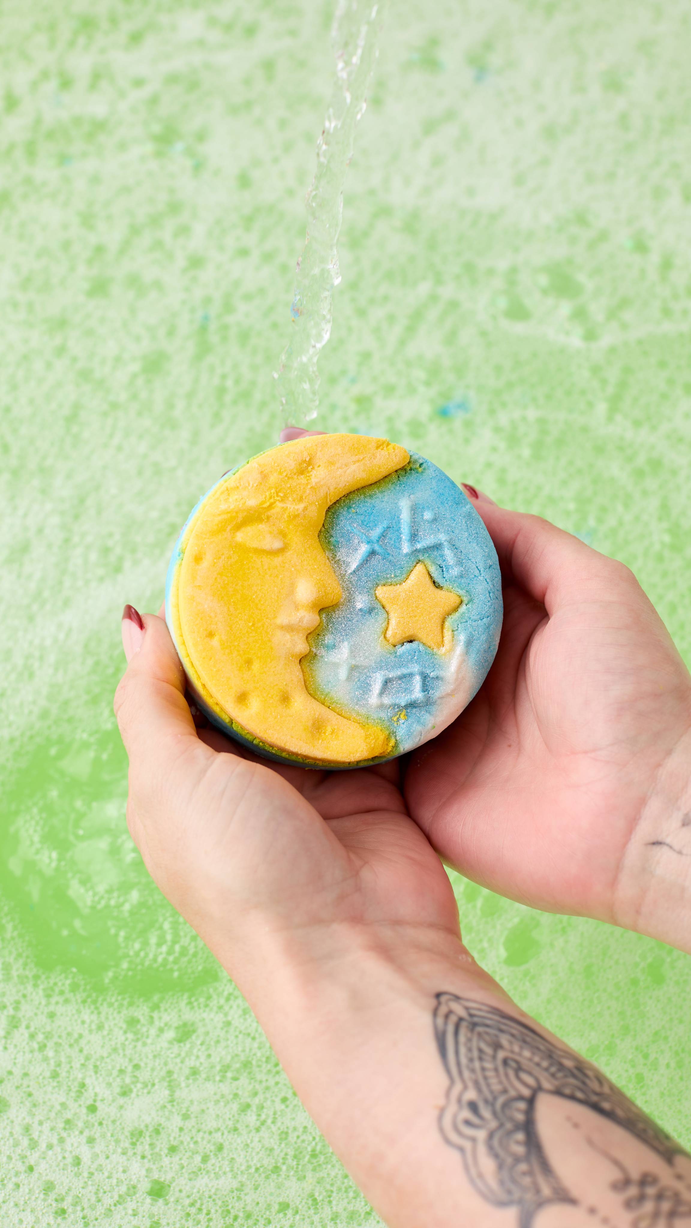 The image shows a hand holding the Christmas Eve bubble bar with a yellow crescent moon and a marbled blue and white sky. It is held under a strong stream of water over a light green, foamy bath.