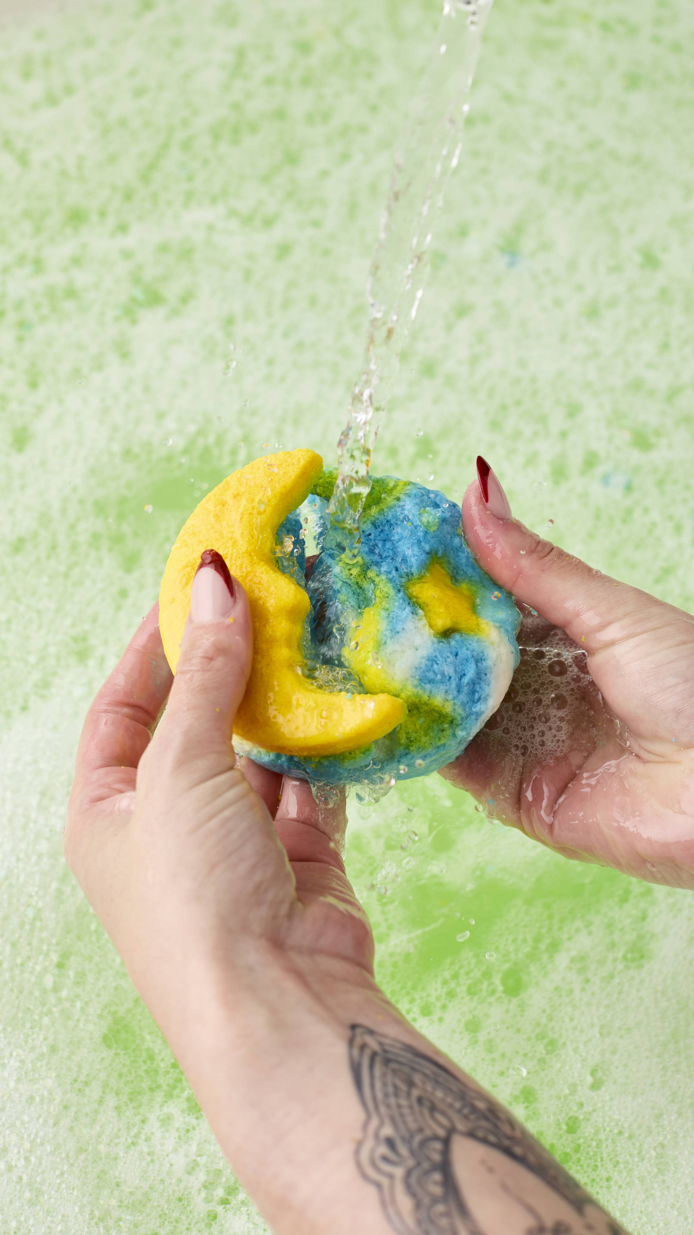 The image shows hands, with long nails, breaking apart the Christmas Eve bubble bar - a round product with a yellow crescent moon and a marbled blue and white sky. It is held under a strong stream of water over a light green, foamy bath.
