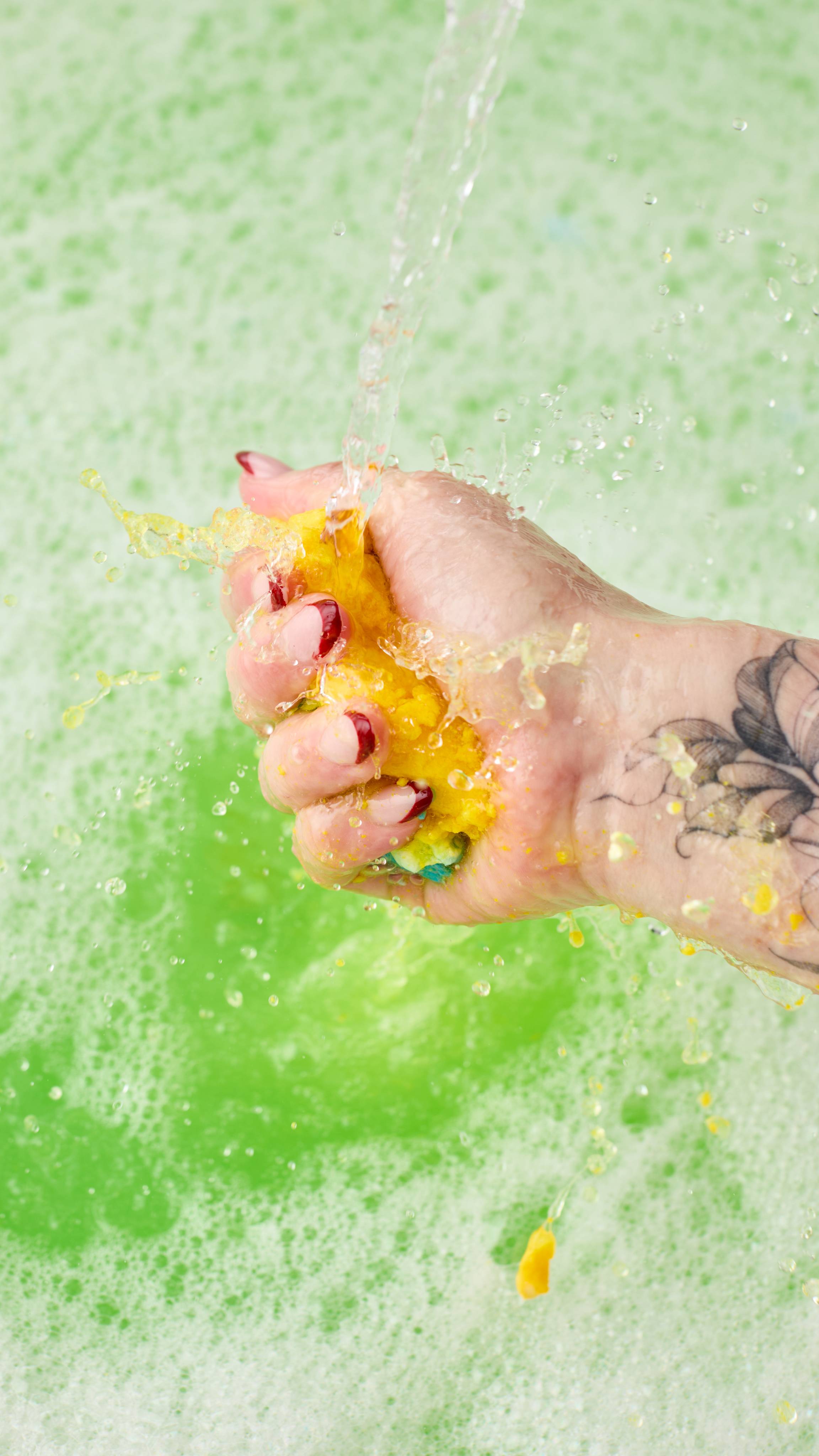 The image shows a hand vigorously squeezing the Christmas Eve bubble bar under a stream of water. Vibrant liquid and water splash off of the product into the vibrant green bath water, creating bubbles and foam.