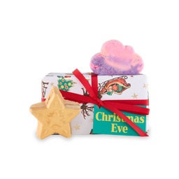 Christmas Eve. A rectangular gift box wrapped in white paper with Christmas tree, reindeer and sleigh illustrations, wrapped in a red ribbon with a green gift tag. Two bath products sit around the gift box.