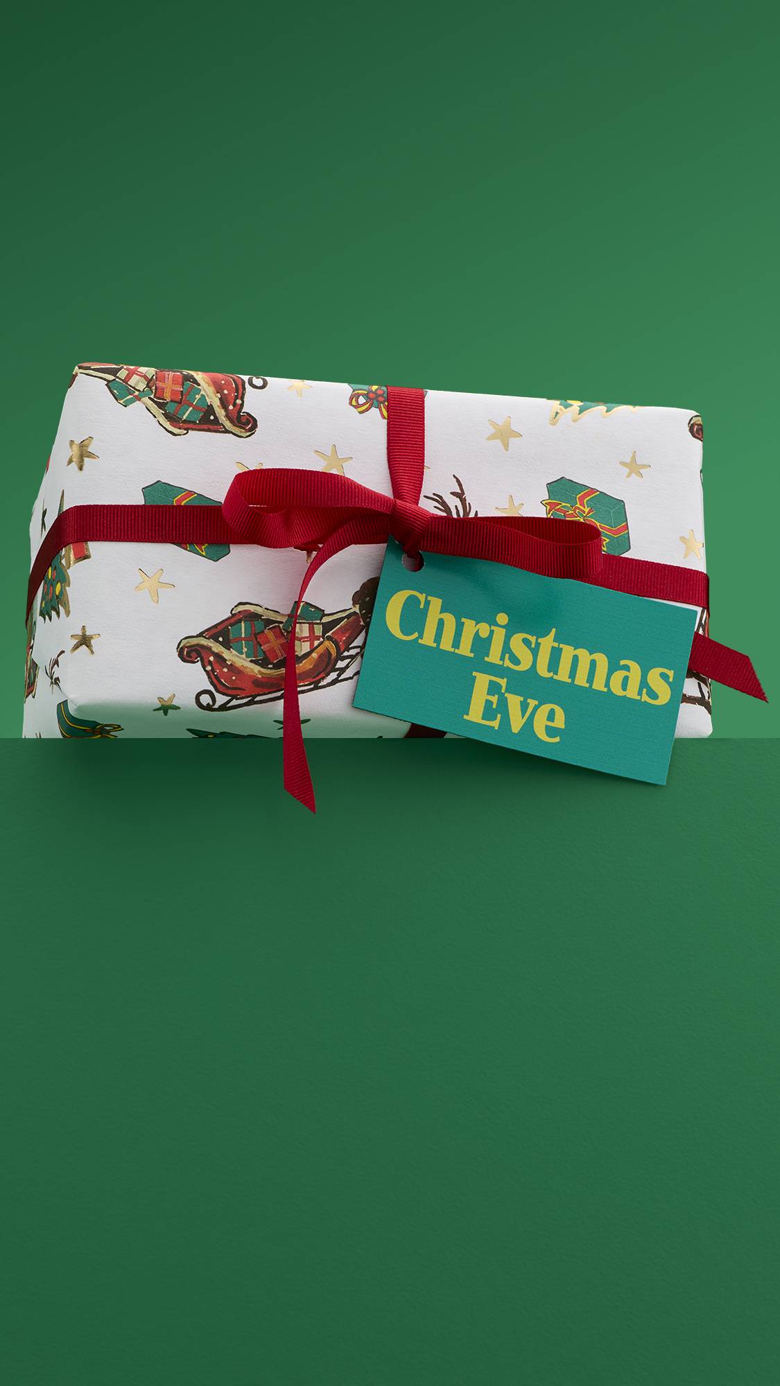 The image shows the vibrant, Christmas Eve gift box, tied with a red ribbon and a green gift tag, sitting on a green platform against a matching green background. 