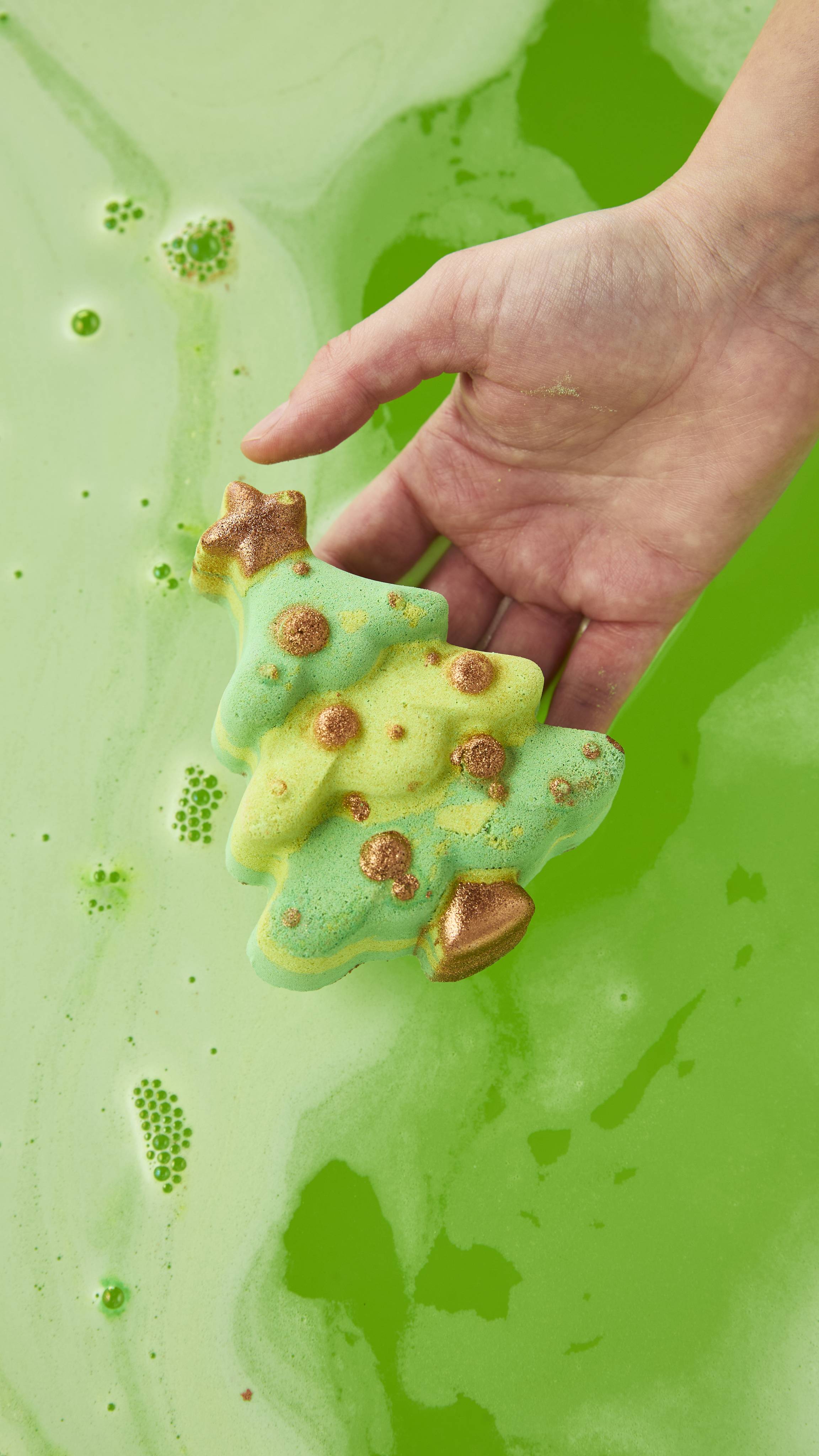 A hand holds a green Christmas tree bath bomb with shimmering gold accents over vivid, lime-green bathwater.