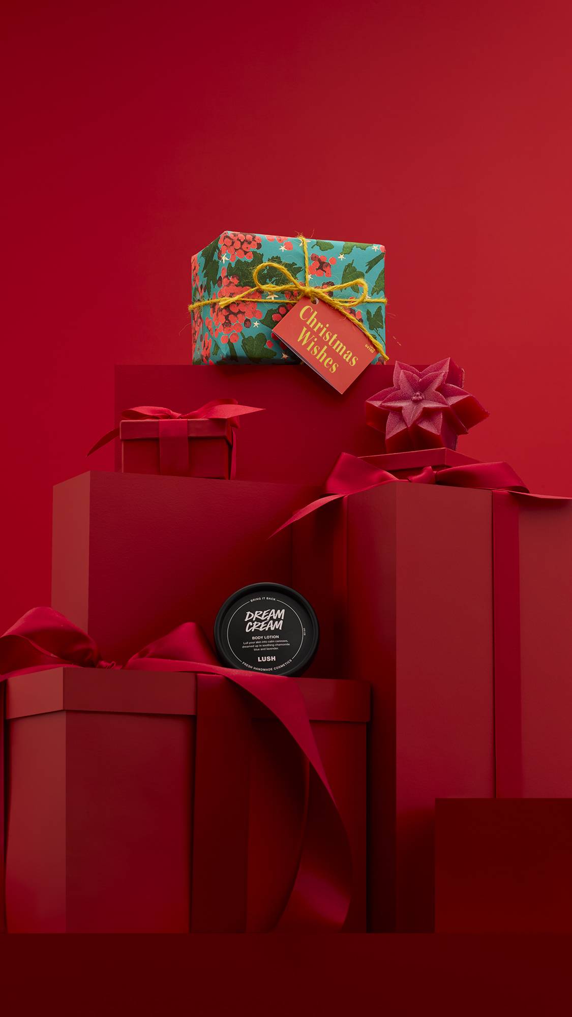 The image shows a large pile of differently-sized wrapped red gifts with ribbons, on a matching red background. The vibrant Christmas Wishes gift box sits on top of the pile, and the two included Lush products sit on other lower red gifts.