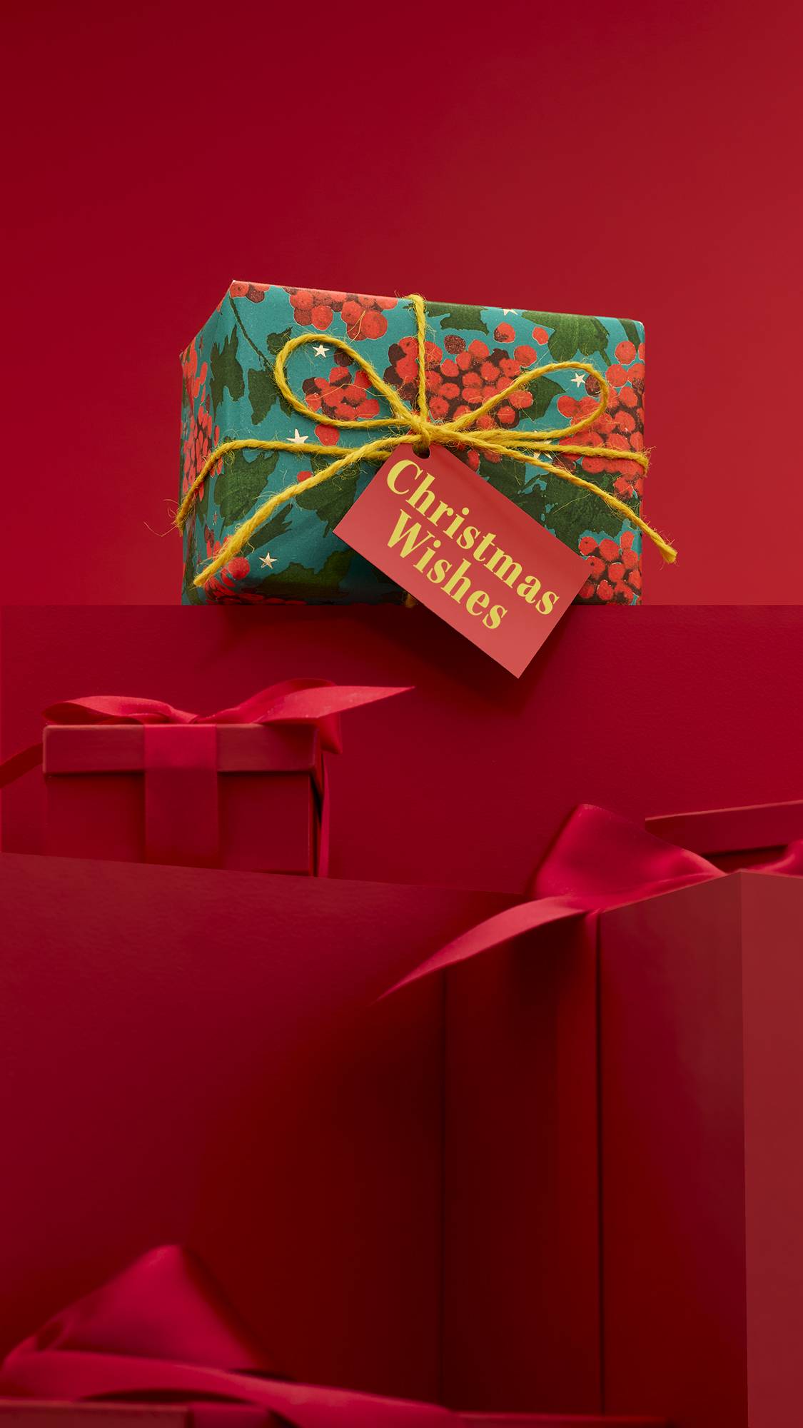 The image shows a large pile of differently-sized wrapped red gifts with ribbons, on a matching red background. The vibrantly colourful Christmas Wishes gift box sits on top of the pile, with green, berry paper, tied with a yellow string. 