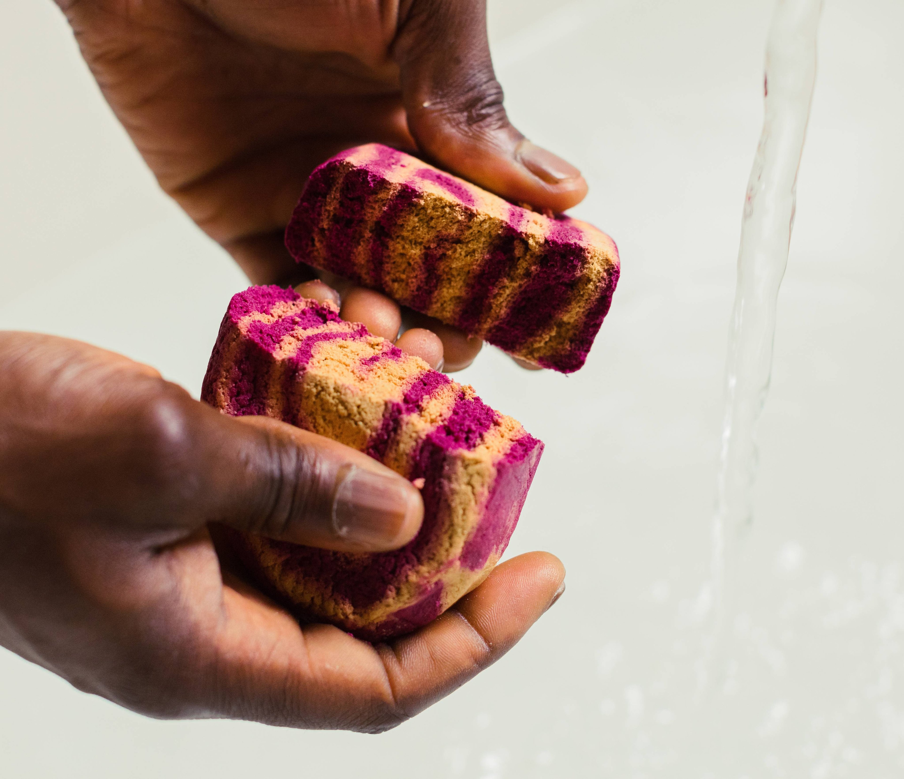Image shows the model breaking the colourful Cinnamon Roll bubble bar in half next to a running tap. 