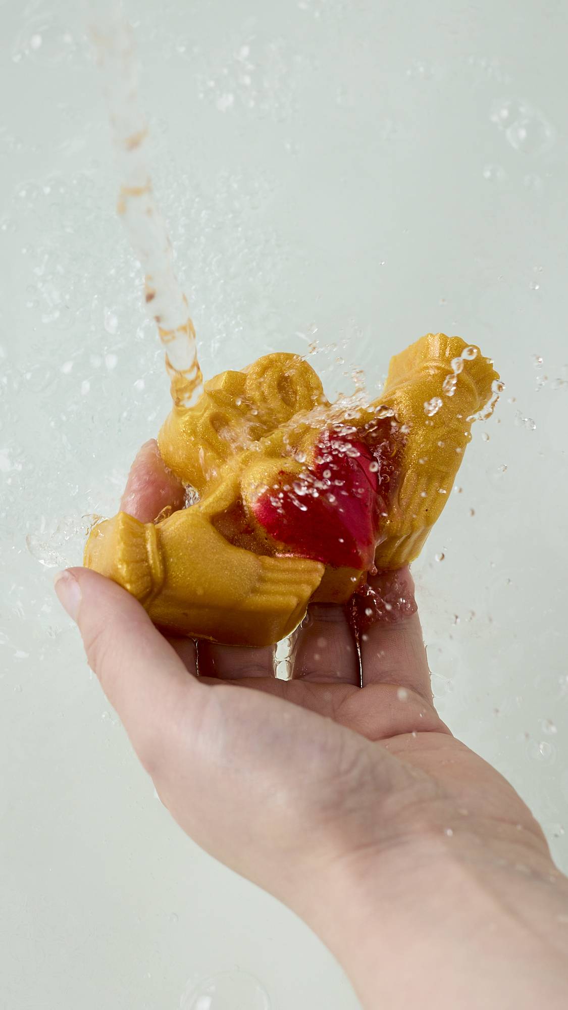 The image shows the model's hand reaching under running tap water above white porcelain. Their hand is holding the traditional Claddagh-shaped soap as droplets splash off the product. 