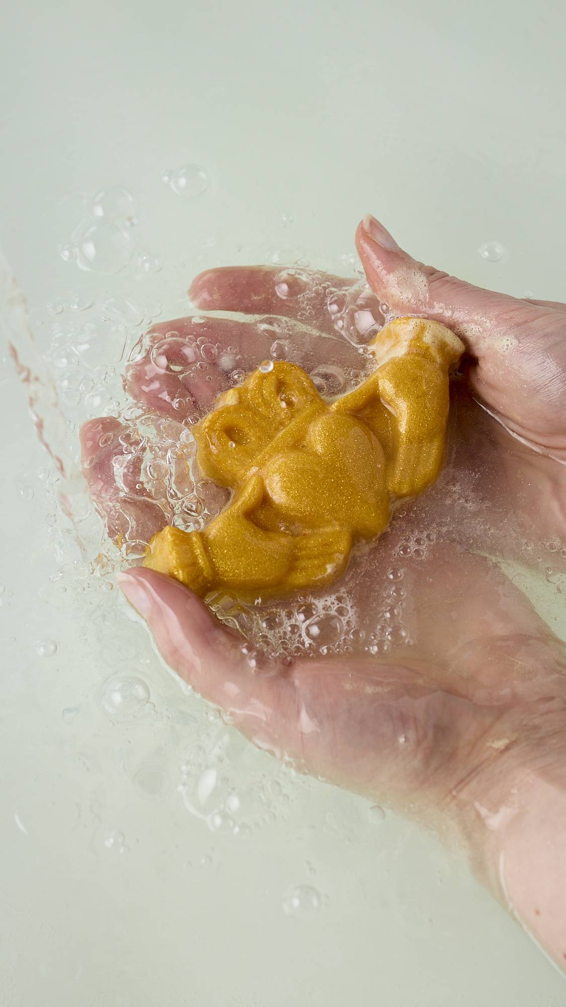 The model's hands are holding the glittering, golden, traditional Irish Claddagh hands, heart and crown shaped soap in their palms after using the product to create a delicate lather over their skin.