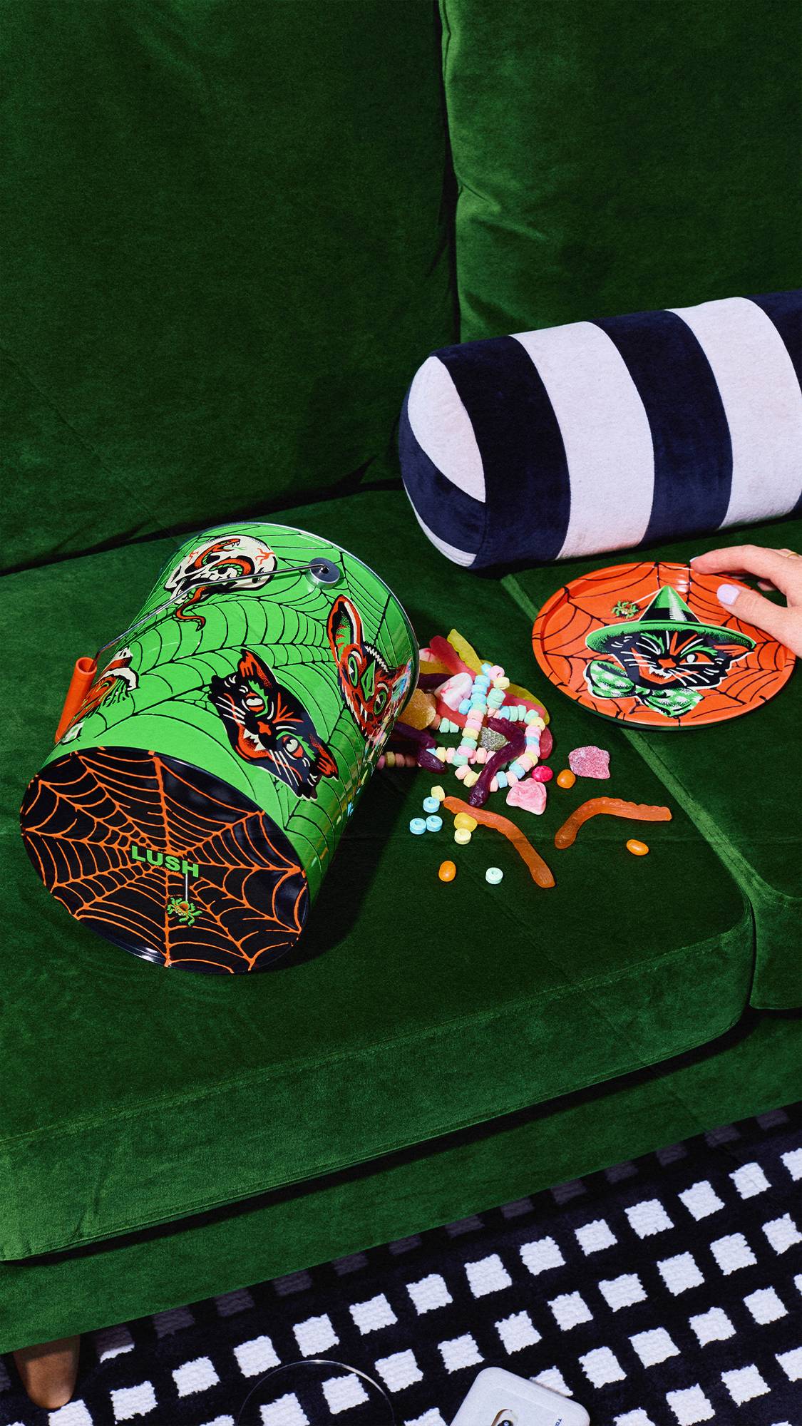A green vintage-style Halloween tin lies on its side on a green velvet couch, with colourful sweets spilling out. An orange lid featuring a black cat design sits nearby next to a striped bolster cushion and a person's hand.