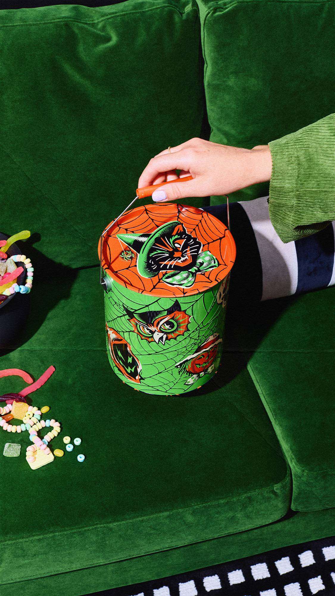 A person's hand holds the handle of a green vintage-style Halloween tin bucket sitting on a green velvet couch. The tin features an owl and pumpkin on a cobweb pattern, with a black cat on the orange lid. Colourful sweets are nearby.