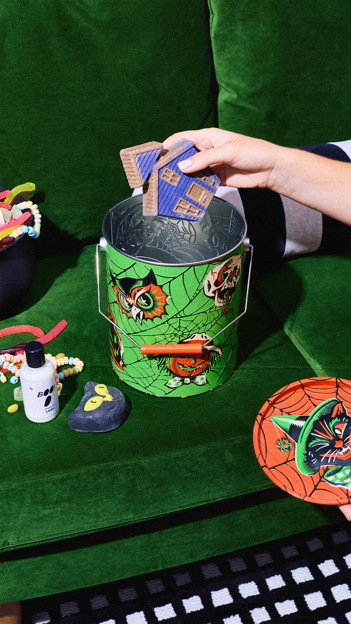 A person's hand places a blue, house-shaped bath bomb into an open green Halloween tin bucket on a green couch. The lid rests nearby, alongside a 'BOO!' shower gel bottle, a black cat bubble bar, and scattered sweets.