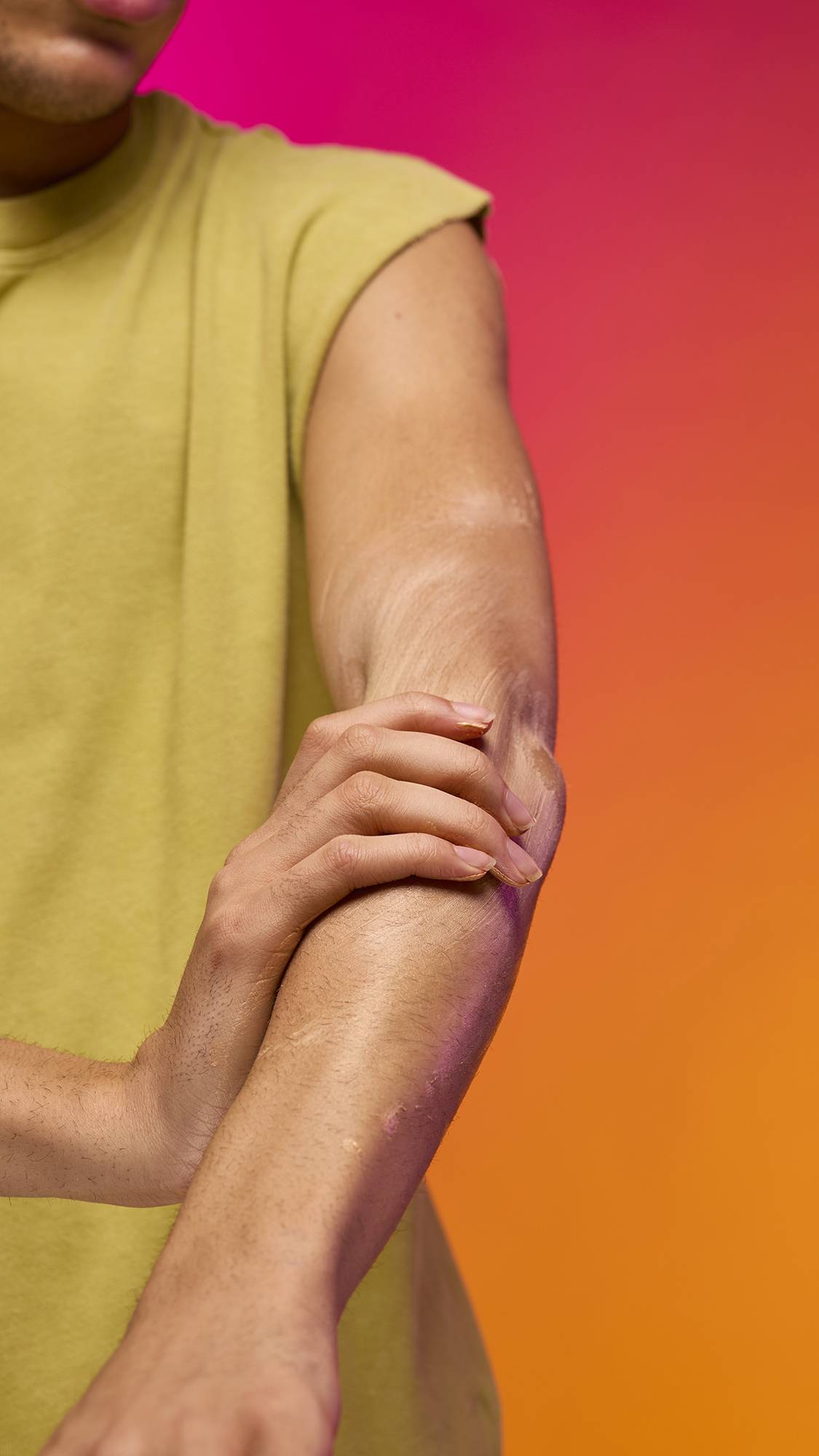 The image shows a portion of the model's torso and their bare arm stretched out as they stand on a pink-to-orange gradient background. They are applying the Cocktail Body lustre up and down their forearm and bicep.