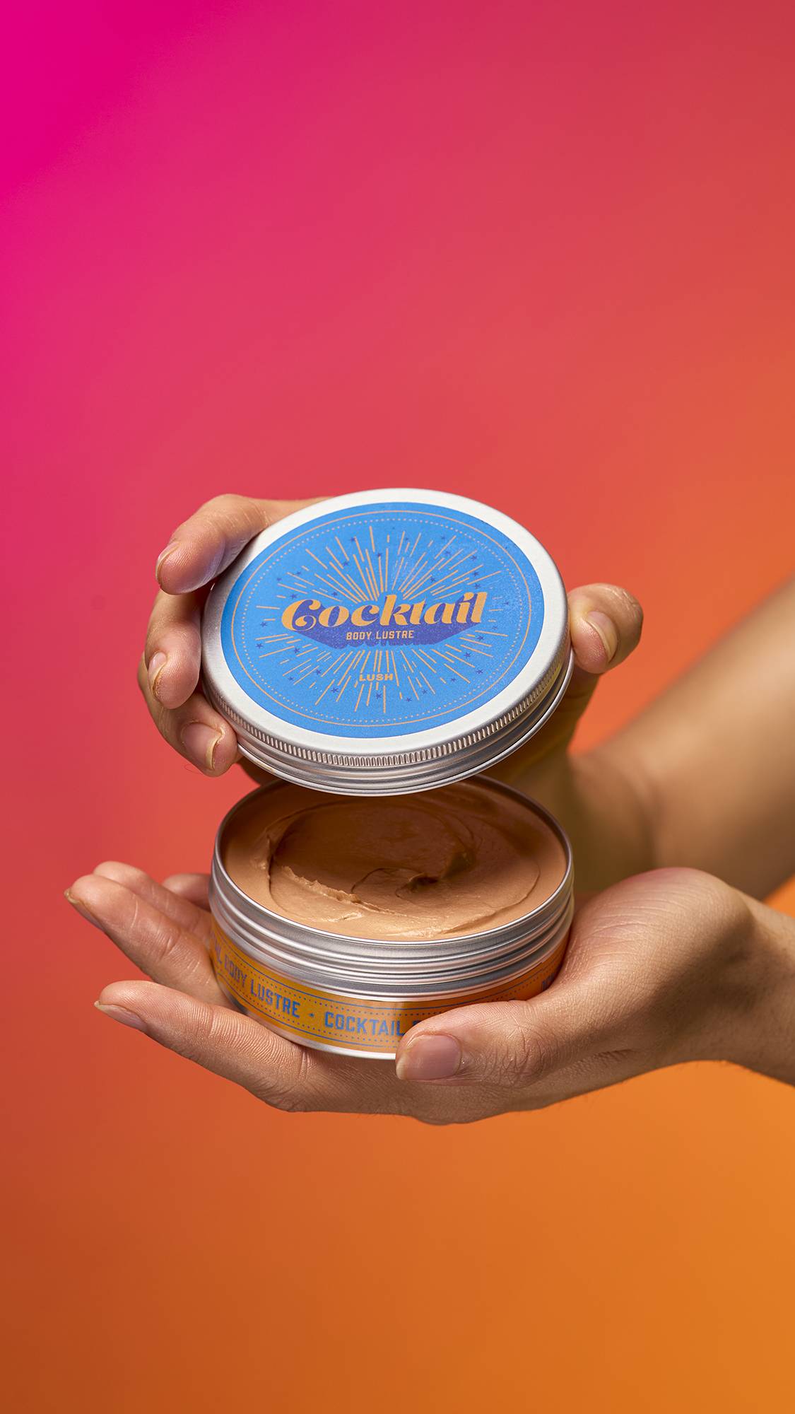 The image shows the model's hands reaching in from the right. They are holding an opened metal tin of the Cocktail body lustre. The packaging has bright stickers and the product appears to have the texture of a smooth, whipped mousse. 