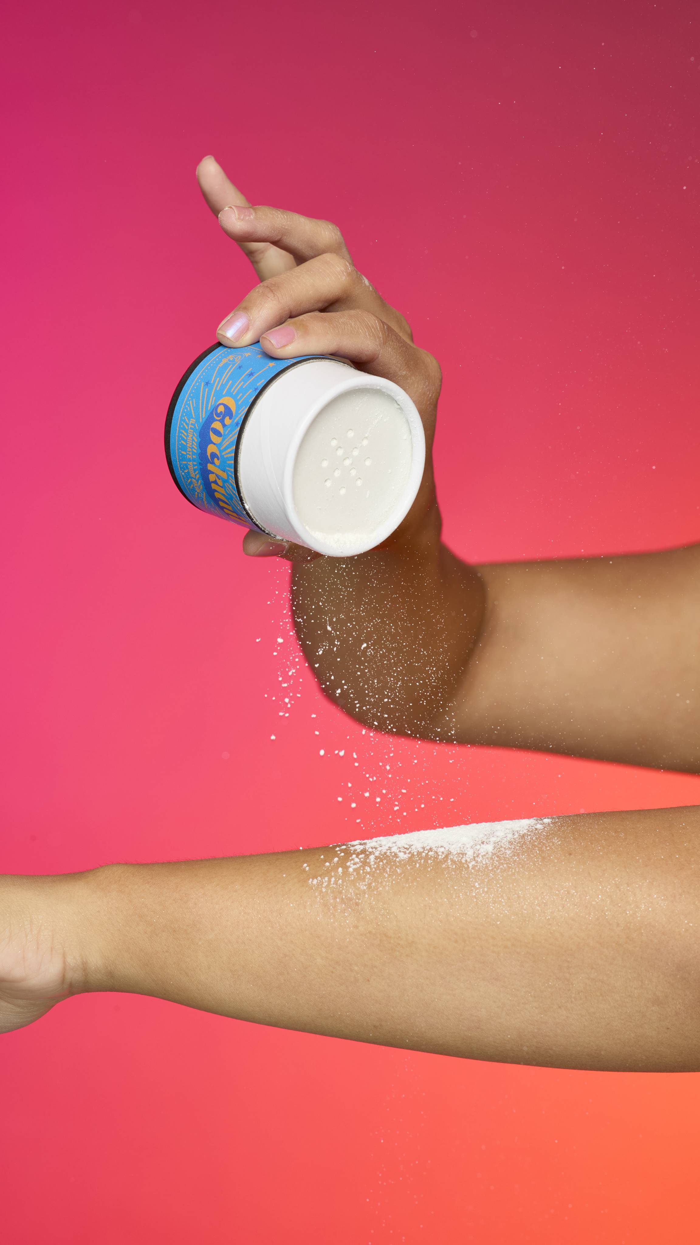 A hand holds a white and blue Lush Cocktail sparkly body powder shaker container, sprinkling the fine powder onto a forearm. The powder is visible in the air and on the skin, and the background is a bright gradient of pink and orange.