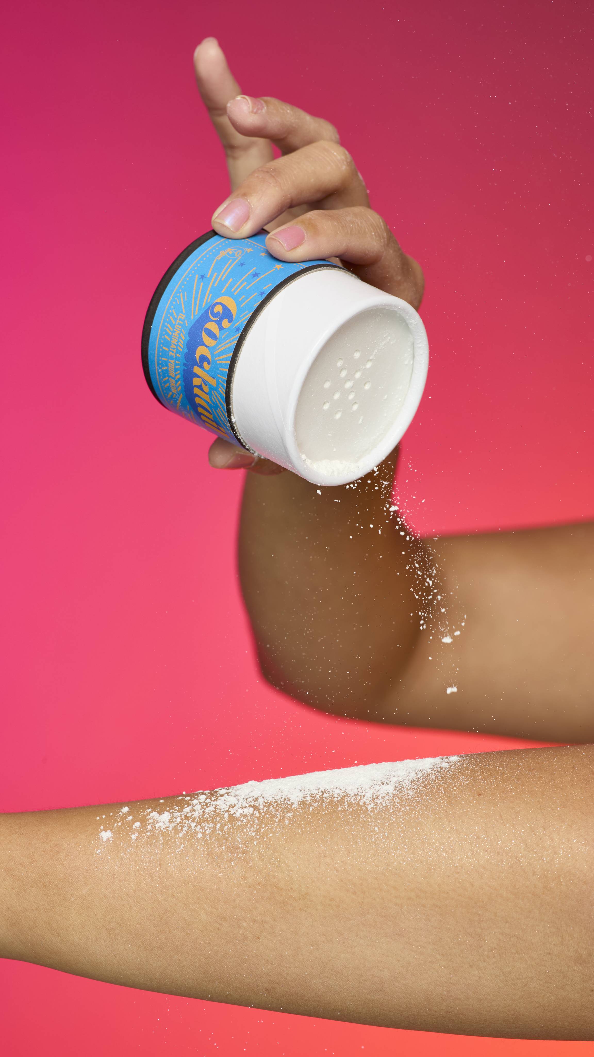 A hand holds a white and blue Lush Cocktail sparkly body powder shaker container, sprinkling the fine powder onto a forearm. The powder is visible in the air and on the skin, and the background is a bright gradient of pink and orange.