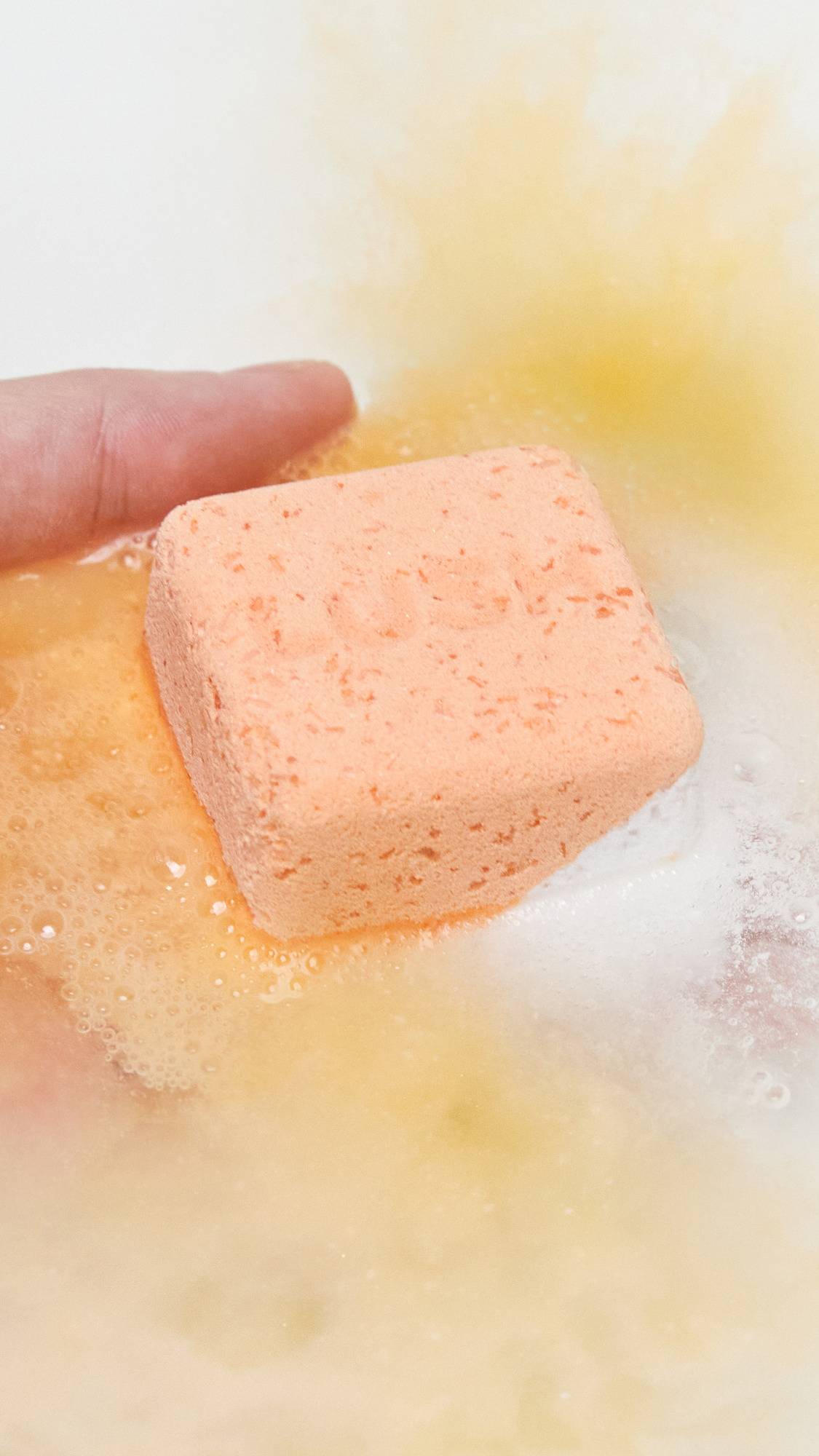 The Cold Water Soother bath bomb is being gently placed into the bath water by hand as it starts dissolving and gives off subtle, peachy foam. 