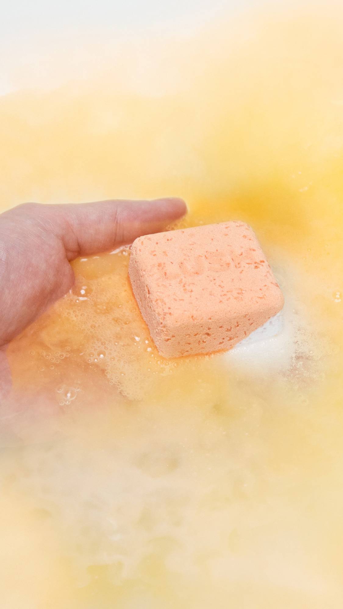 The Cold Water Soother bath bomb is being gently placed into the bath water by hand as it starts dissolving and gives off subtle, peachy foam. 