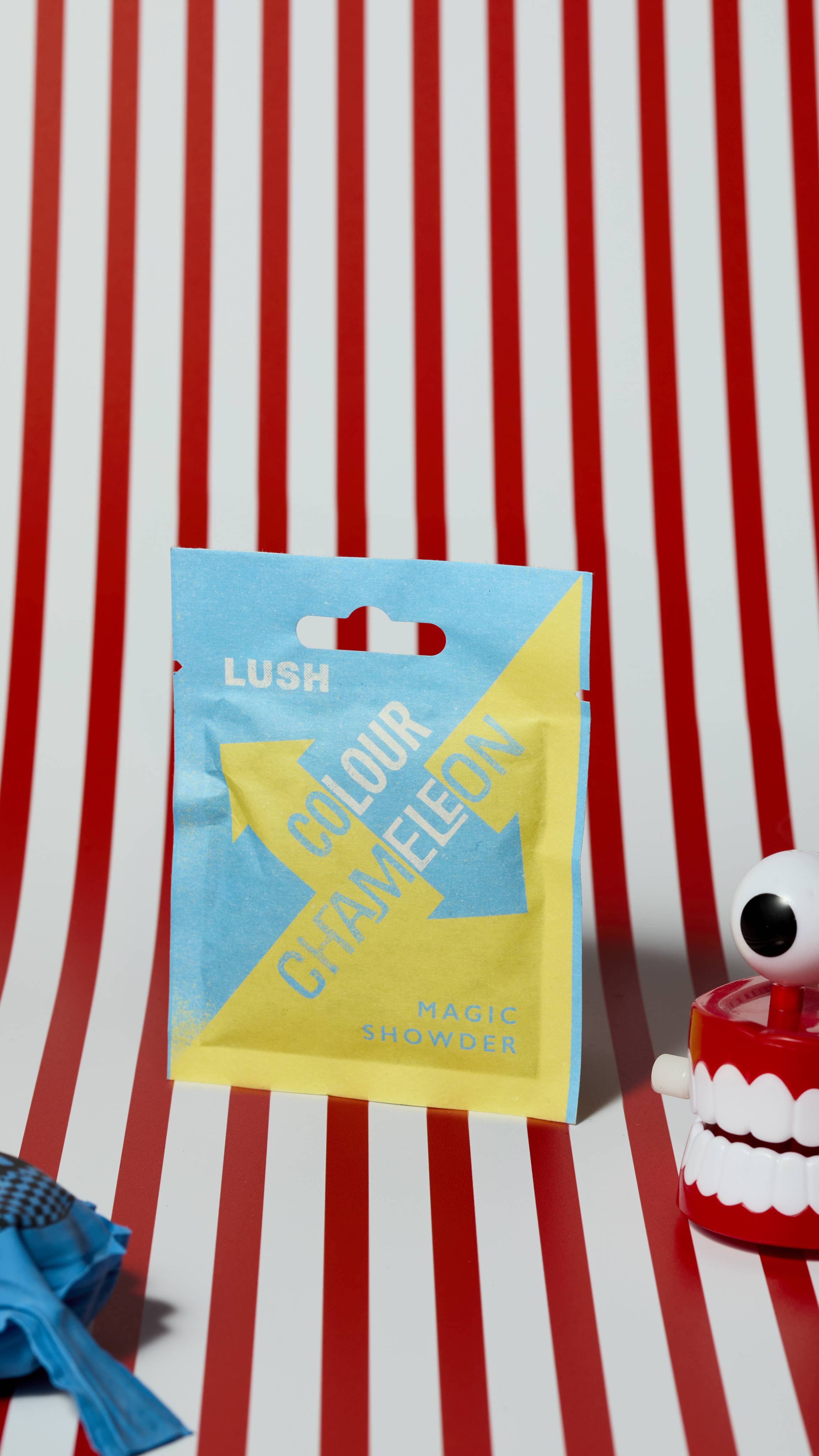 The image shows the blue and yellow sealed paper sachet of Colour Chameleon shower on a red and white striped background. There is a whoopie cushion and wind-up chattering teeth that are just visible.
