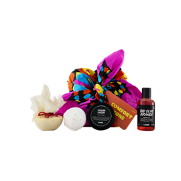 A bright pink knot-wrapped "COMFORT ZONE" gift set sits alongside Lush products, including a white muslin-covered bath melt, a white bath bomb, a black tub of Dream Cream, and a bottle of The Olive Branch shower gel.
