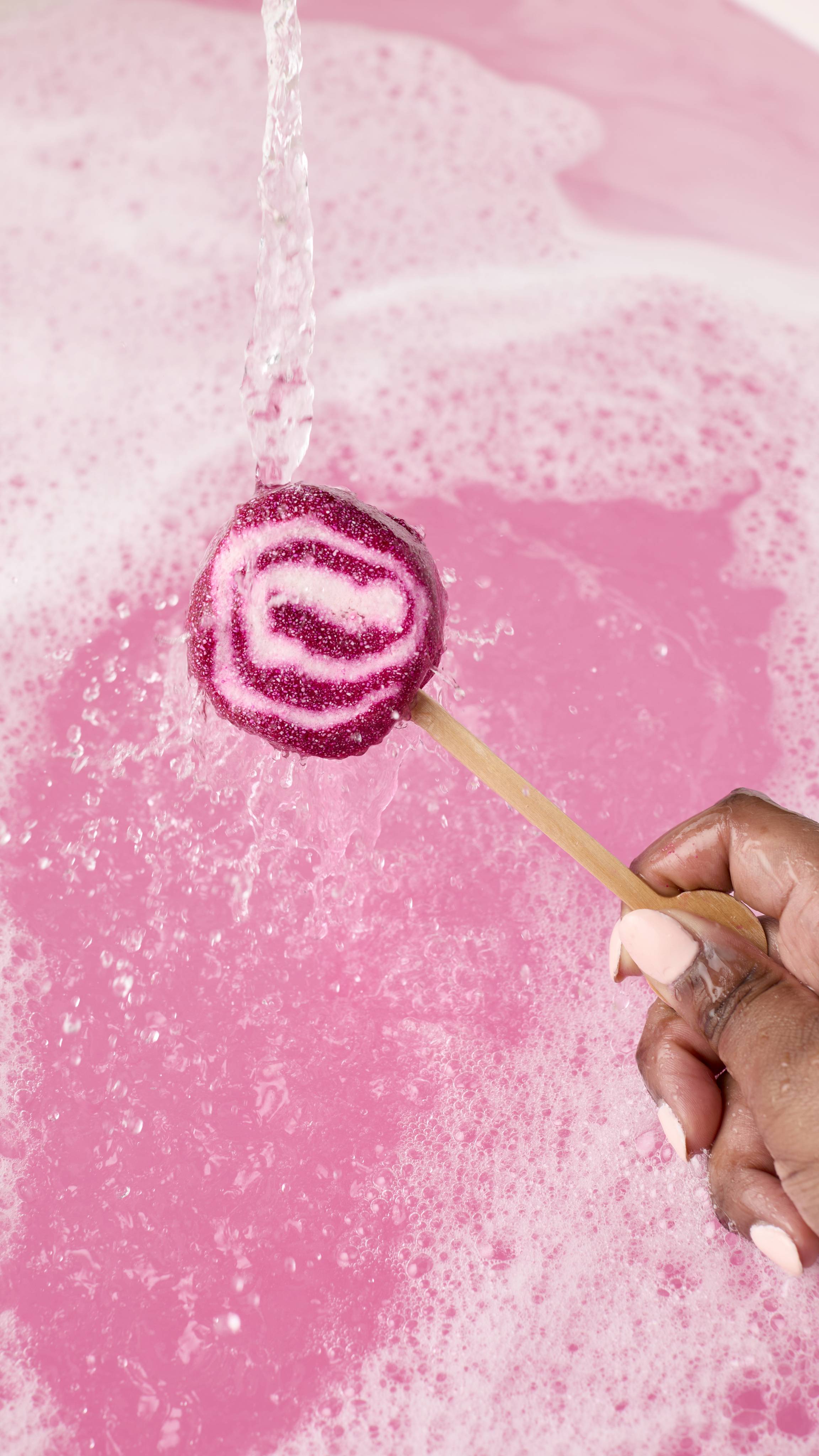 A hand holds a magenta and white swirled Comforter mini bubble wand by its wooden handle. Water pours over the wand into a bright pink bath filled with white bubbles.