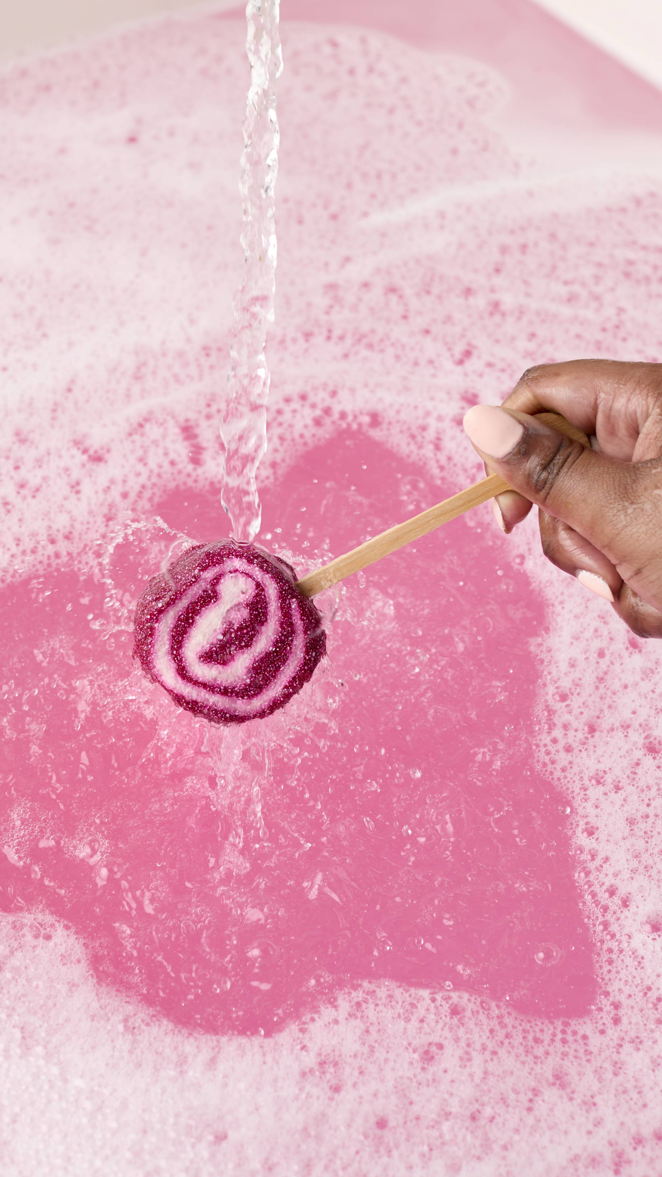 A hand holds a magenta and white swirled Comforter mini bubble wand under a stream of running water. The wand is being held over a bright pink bath filled with white bubbles.
