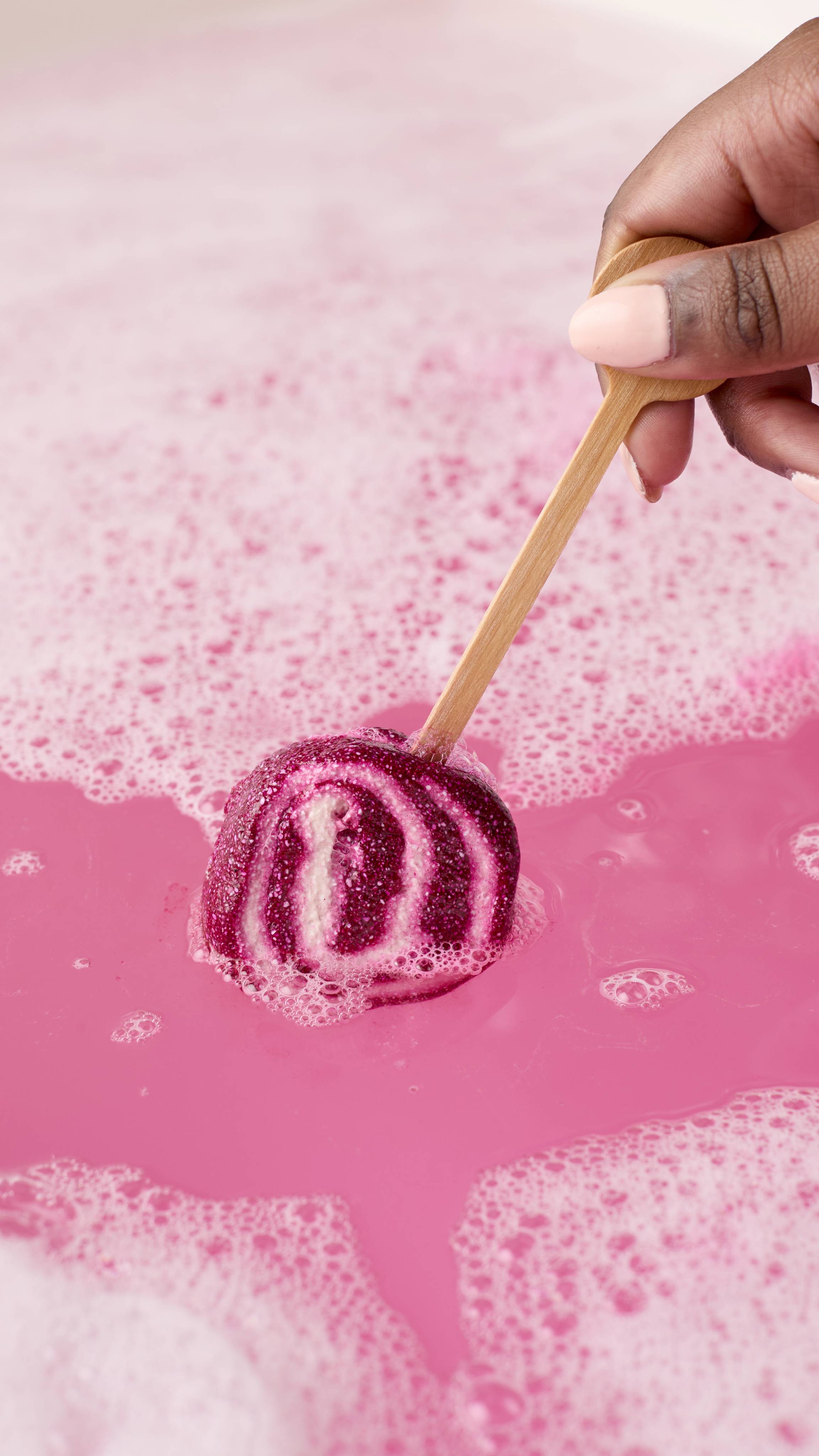A hand holds a magenta and white swirled Comforter mini bubble wand by its wooden handle. The wand is being swirled into a bright pink bath covered in white, frothy bubbles.